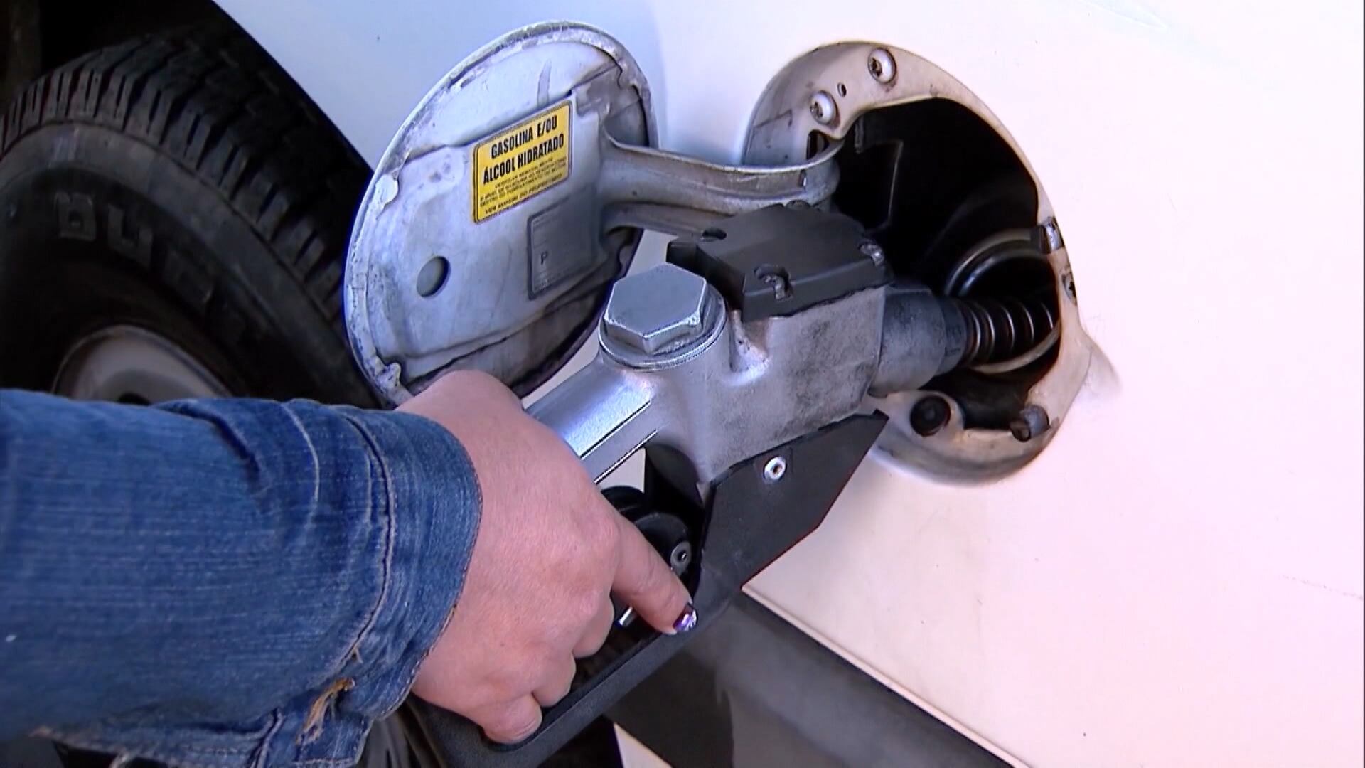 Pessoa abastece carro branco em posto de combustível. A mão segura o bico da bomba encaixado no tanque.