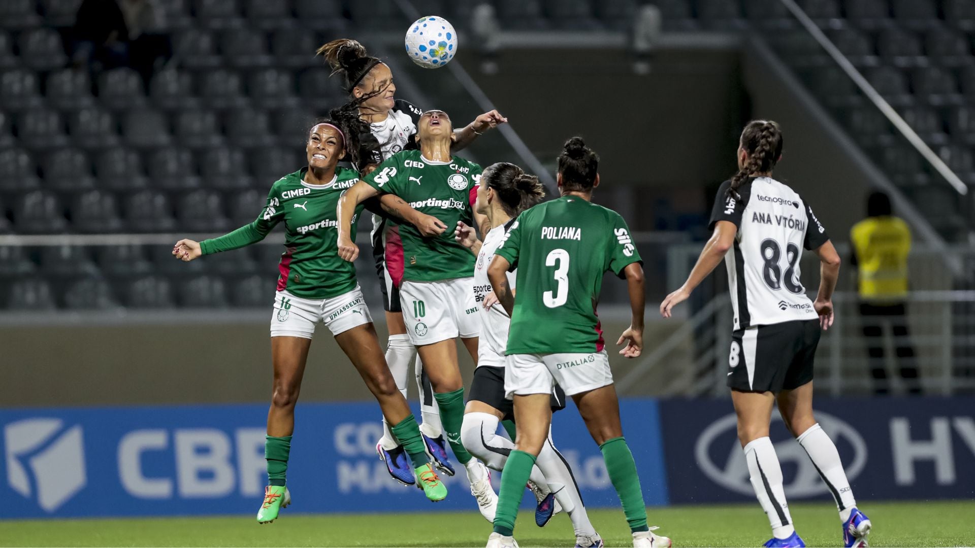 Partida de futebol feminino mostra várias jogadoras saltando para disputar bola aérea. Atletas de uniforme verde, enfrentam adversárias de uniforme branco e preto.