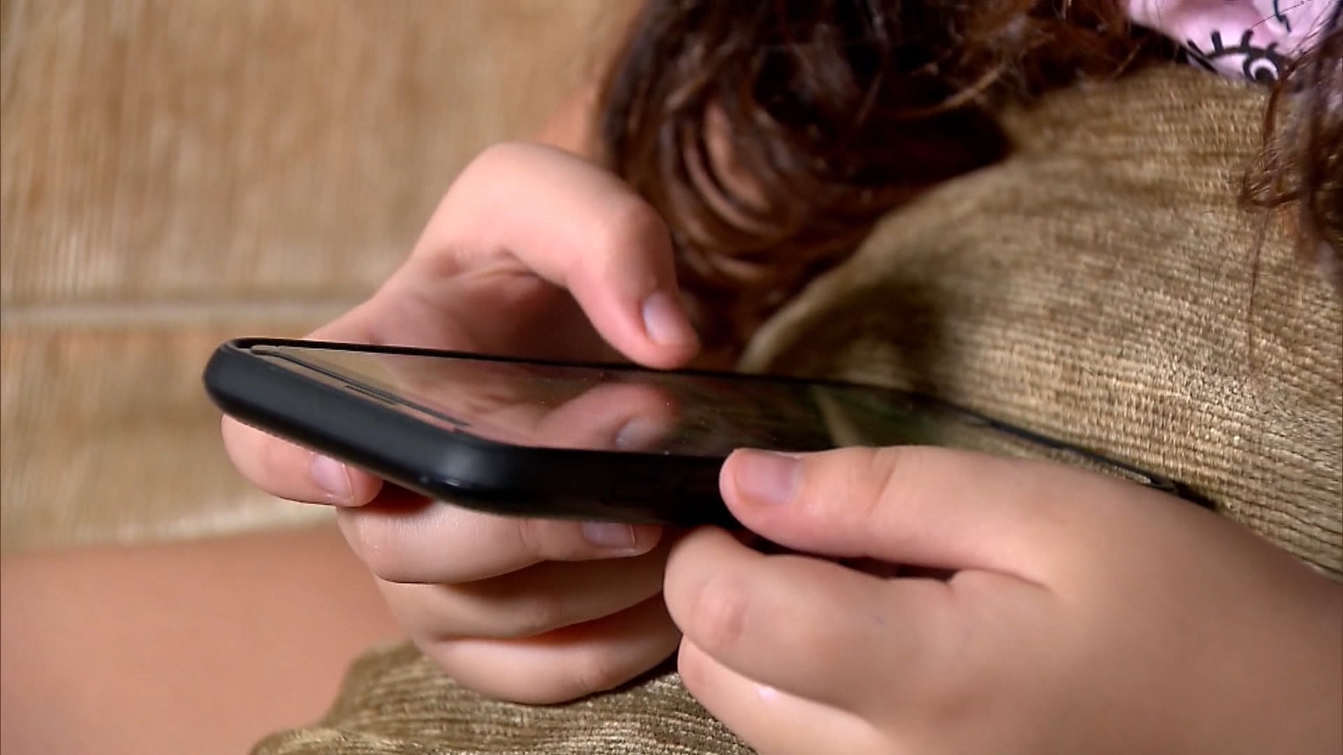 Pessoa segura celular com as duas mãos, interagindo com a tela. Fundo mostra tecido texturizado, parecido com sofá, além de parte da roupa e cabelo da pessoa.
