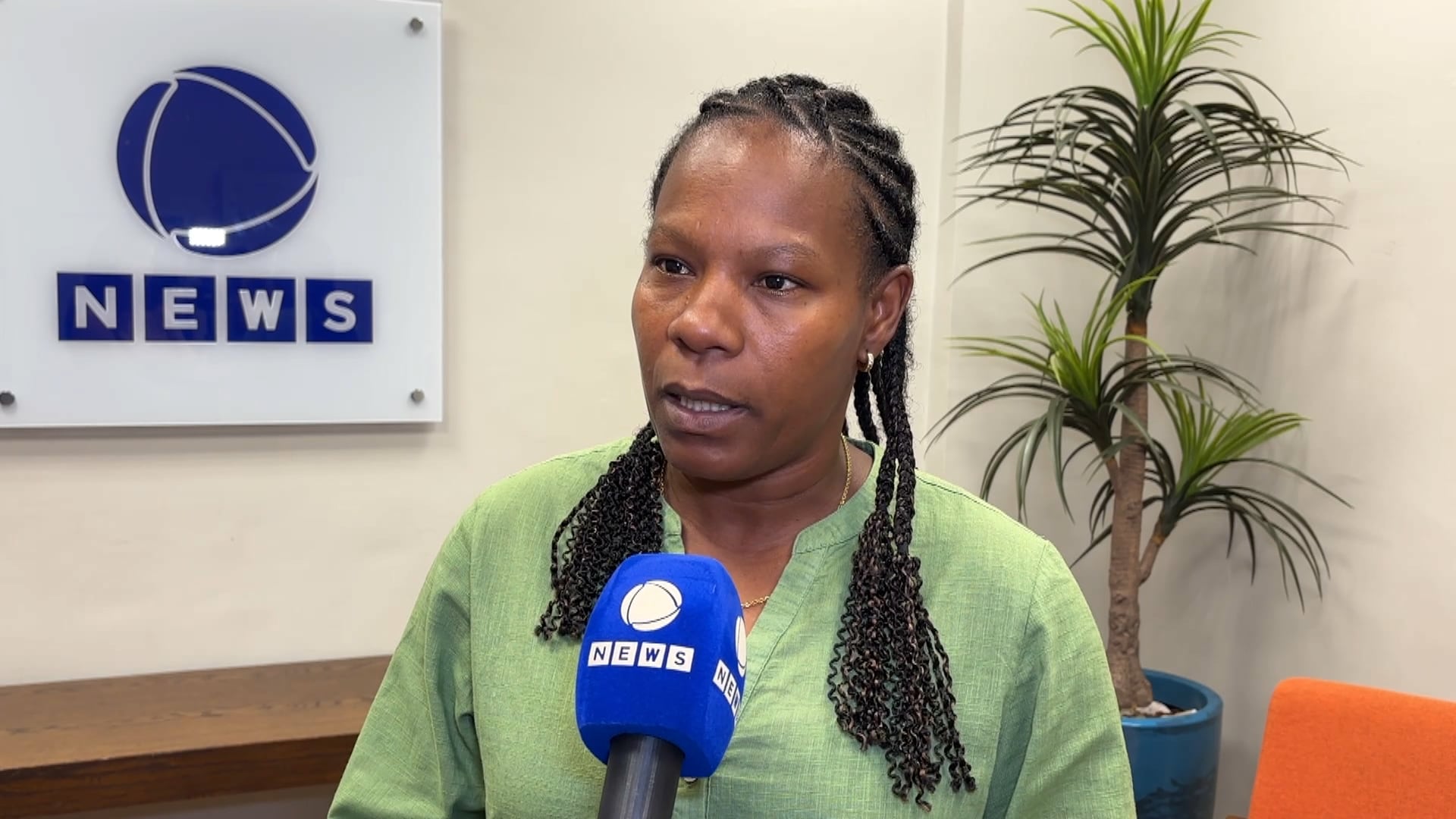 Mulher de tranças e blusa verde fala em microfone azul com logotipo “NEWS”. Ao fundo, parede branca exibe placa também com a palavra “NEWS” e desenho de globo.