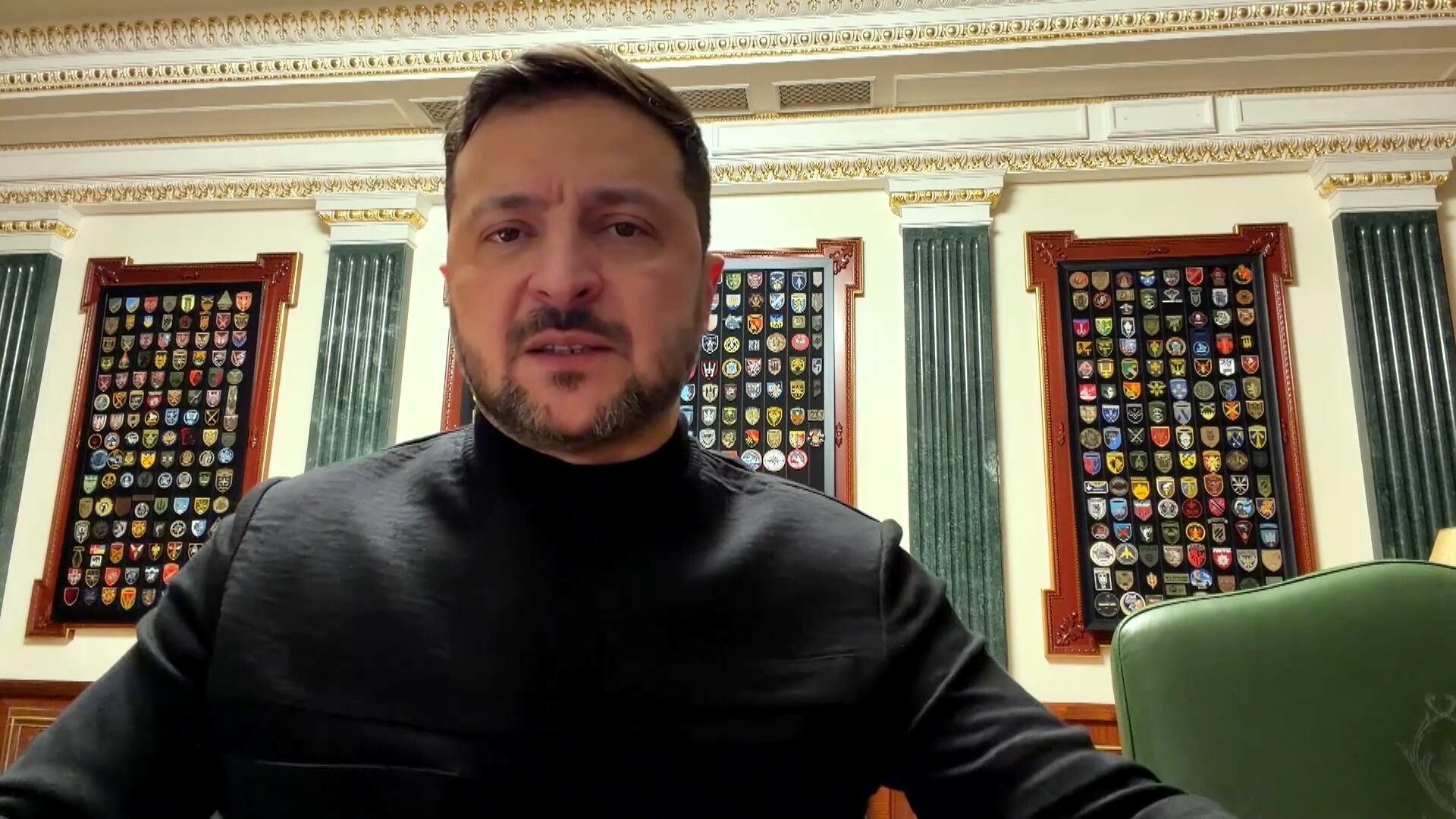 Volodymyr Zelesnky veste uma roupa preta e está sentada em uma sala com paredes decoradas por painéis contendo diversos brasões coloridos