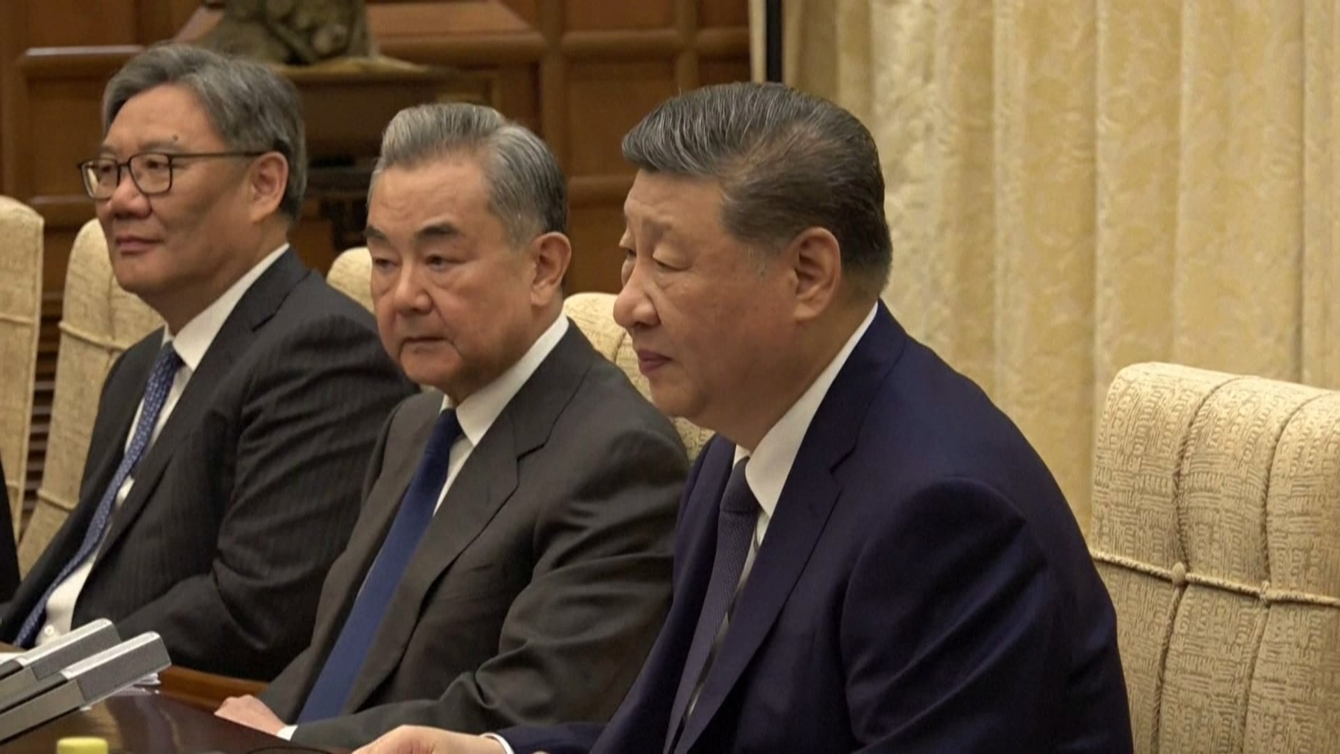 Xi Jinping veste terno azul escuro; dois homens sentados ao lado dele em uma sala de reuniões, vestindo trajes formais e posicionados diante de uma mesa