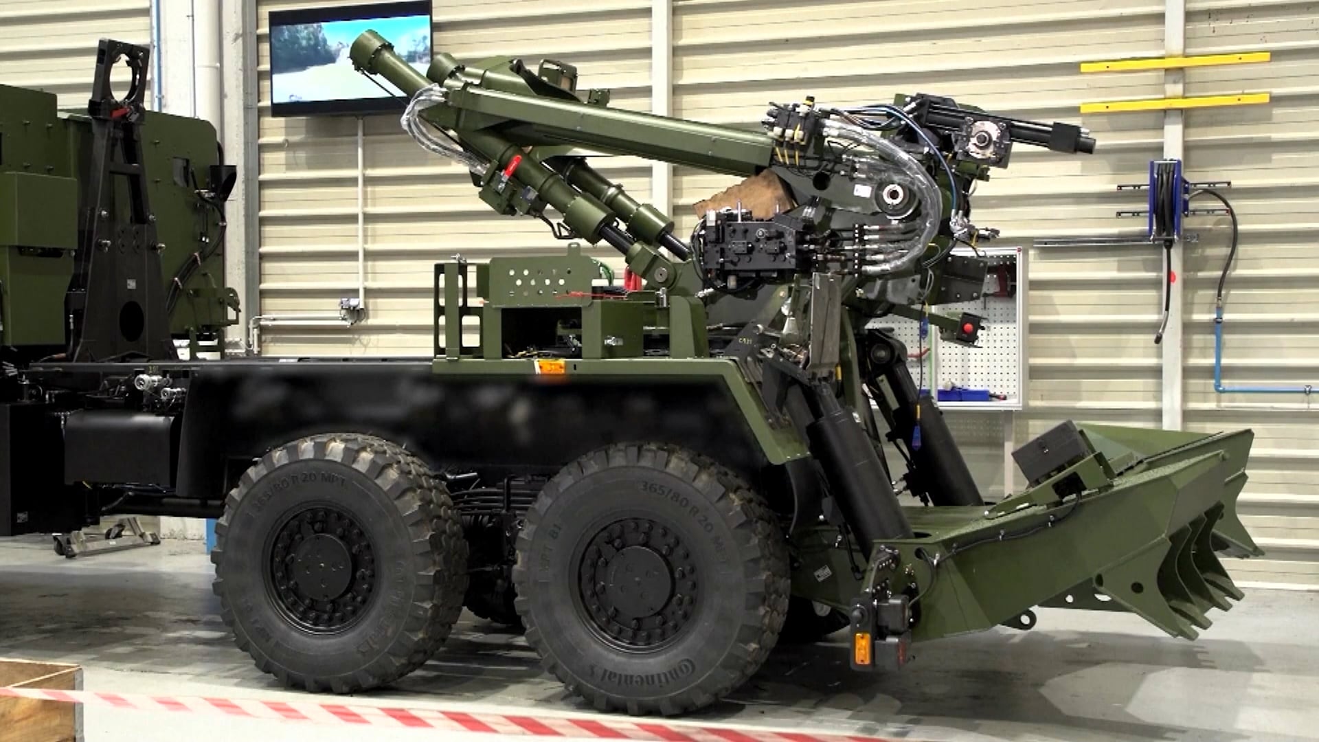 Veículo militar com seis rodas, equipado com braço mecânico articulado, ferramentas hidráulicas e componentes metálicos expostos, estacionado dentro de galpão industrial