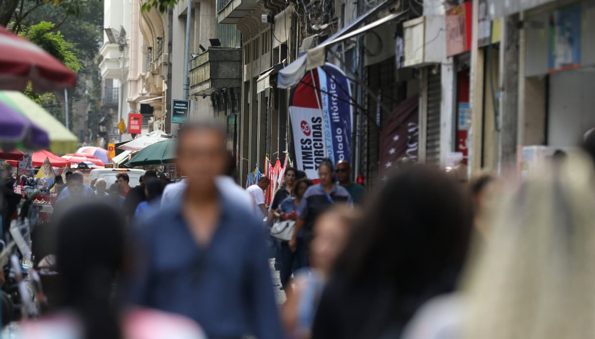 Rua movimentada com várias pessoas caminhando. As que estão visíveis, estão com o rosto embaçado