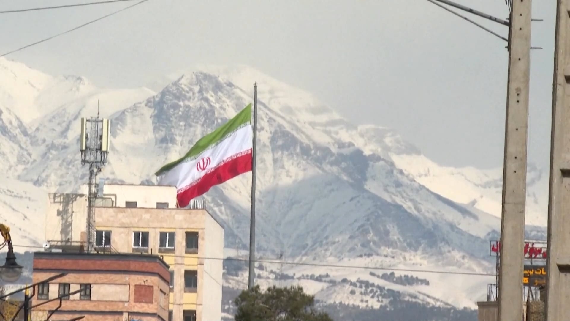 Bandeira do Irã hasteada em um mastro alto no centro de um ambiente urbano, com edifícios e torres de comunicação em primeiro plano e montanhas cobertas de neve ao fundo