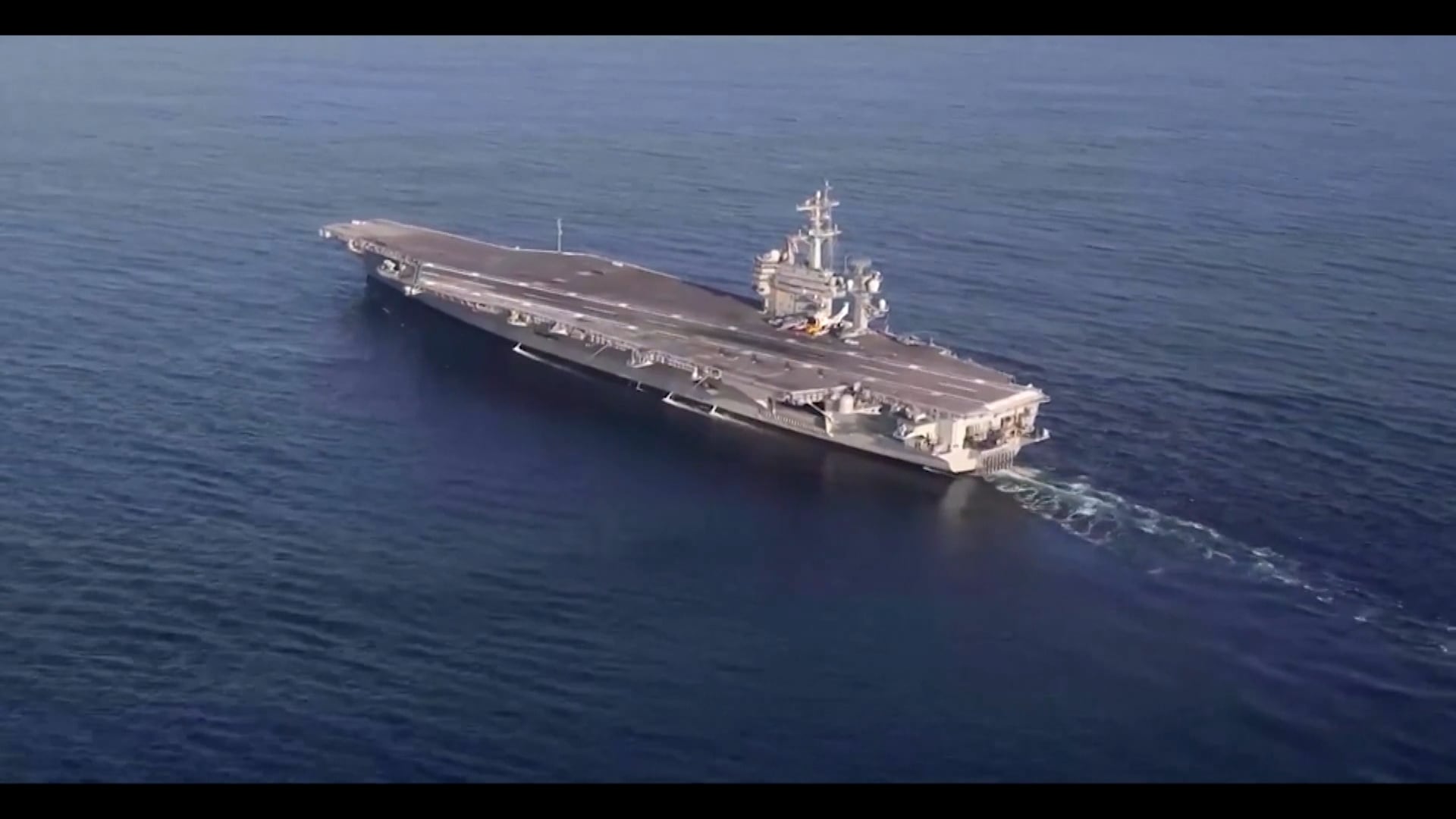 Porta‑aviões navegando em mar aberto, visto de cima, com ampla pista de pouso e estruturas de comando ao centro da embarcação