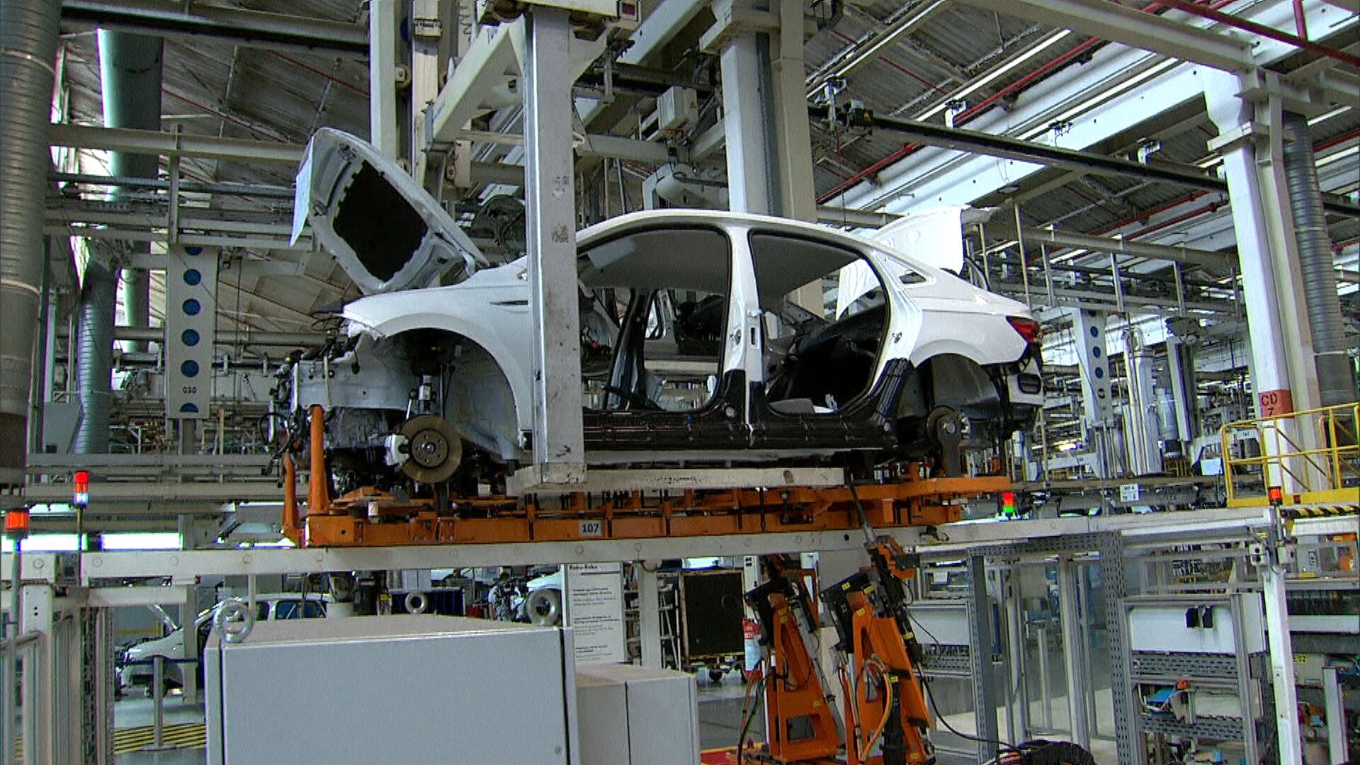 A imagem mostra o interior de uma fábrica de automóveis, com uma carroceria branca parcialmente montada suspensa por uma estrutura metálica. A carroceria está sem portas, rodas e componentes internos. Equipamentos robóticos e braços mecânicos cercam a área, enquanto tubulações, trilhos e suportes metálicos ocupam o teto e o fundo do galpão. O ambiente é iluminado por luz branca industrial.