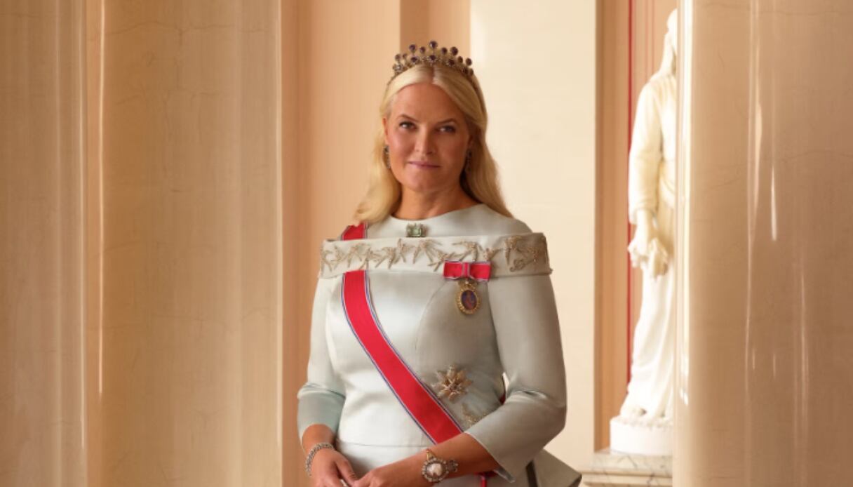 Princesa Herdeira da Noruega com um vestido branco e uma faixa vermelha atravessando o peito. Ela usa uma coroa e tem os cabelos soltos