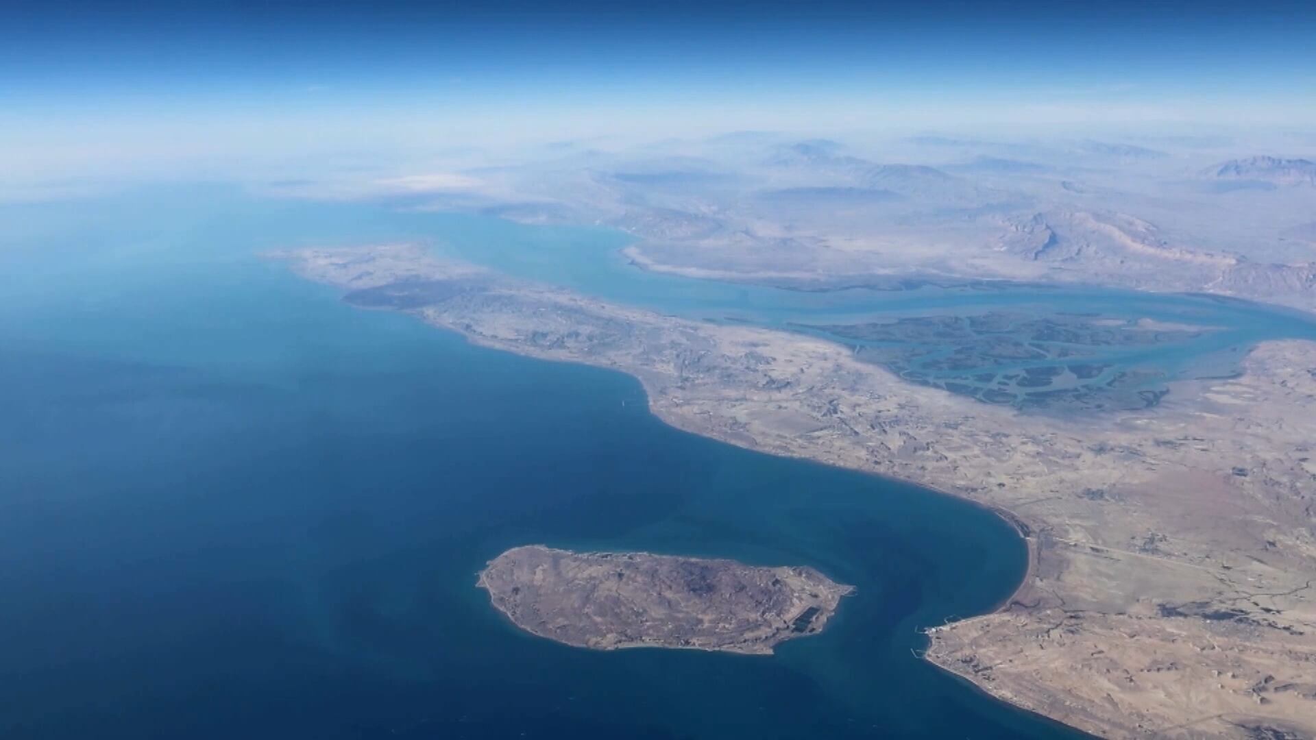 Fotografia aérea do Estreito de Ormuz, no Irã, onde uma grande faixa de costa árida é cercada pelo mar. No centro, há uma ilha rochosa de formato irregular
