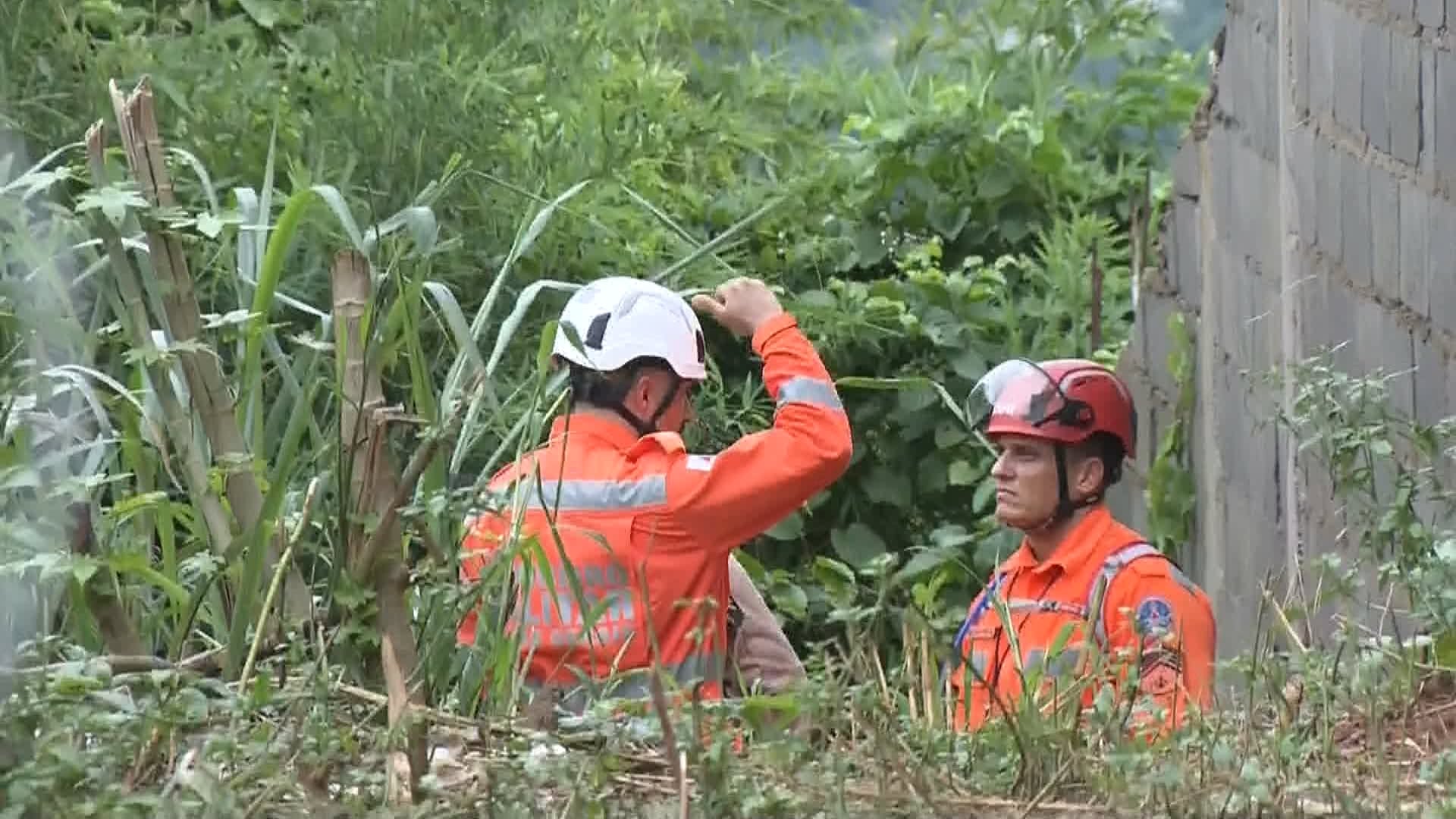 Dois socorristas, usando capacetes e uniformes laranja, conversam em uma área com vegetação alta e densa. Uma parede de tijolos aparece à direita, e o terreno ao redor é irregular, com plantas e galhos.