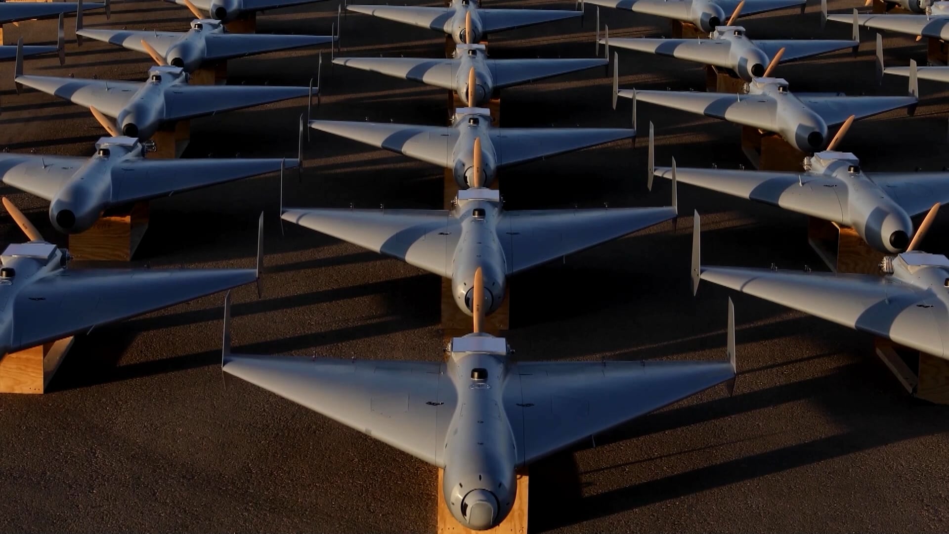Vários drones idênticos estão alinhados em formação sobre o chão. Cada um tem asas retas e hélice traseira. Eles estão apoiados em suportes de madeira.