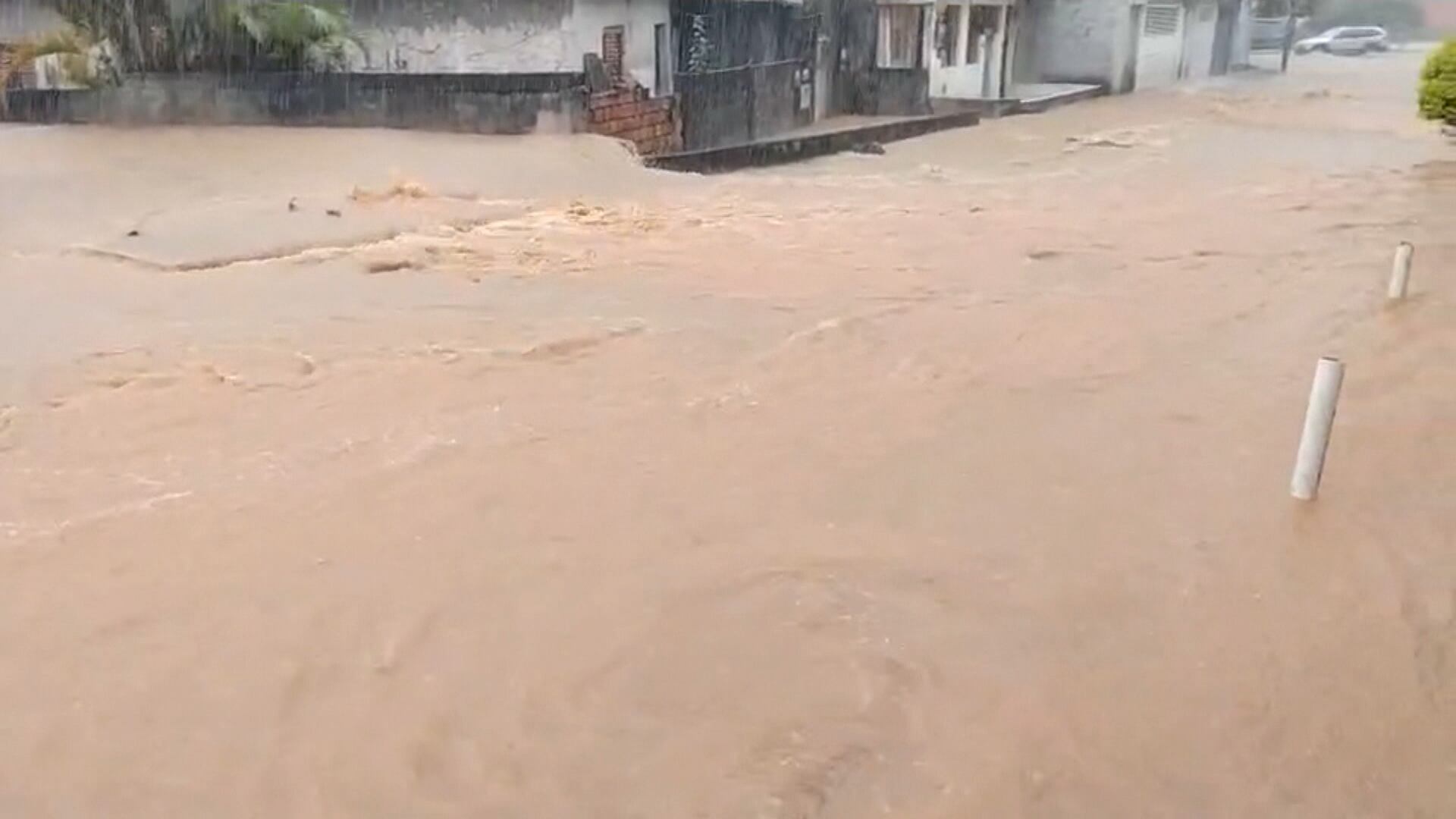 Rua é tomada por uma forte enxurrada de água barrenta, que chega próximo às calçadas das casas. Ao fundo, é possível ver um carro com as rodas cobertas e a chuva caindo com força.