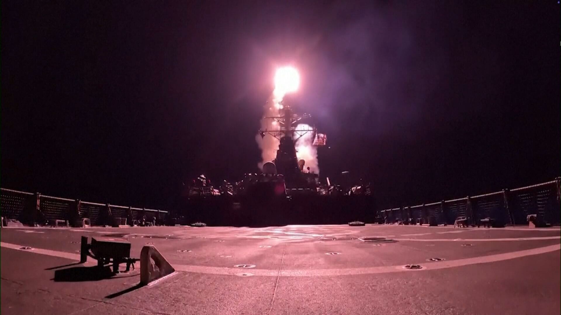 Cena noturna em navio de guerra mostra o lançamento vertical de um míssil. A chama e a fumaça iluminam o convés, onde há grades de segurança e equipamentos.