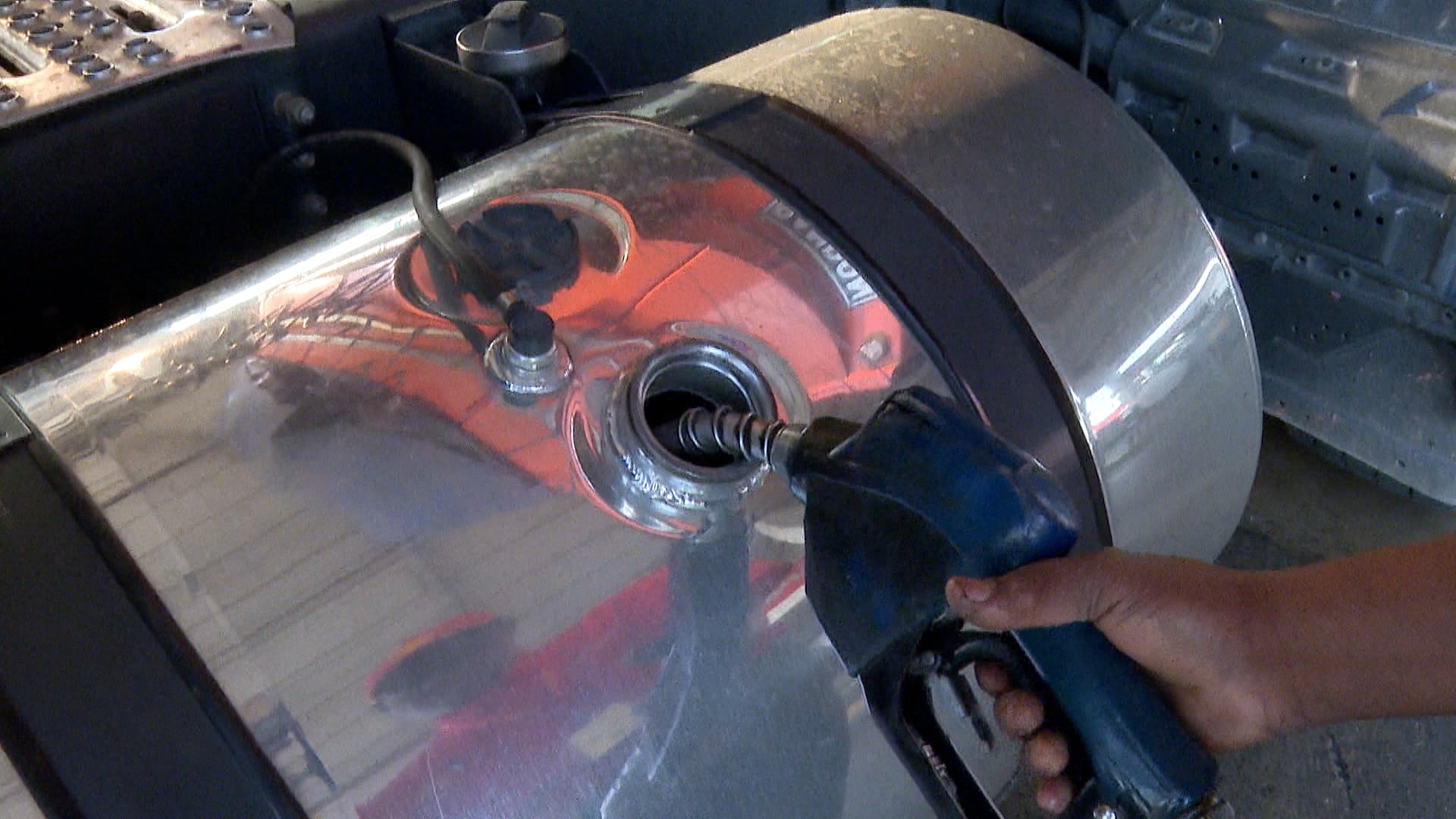 Pessoa segura bico de combustível azul encaixado em tanque metálico cilíndrico e brilhante de caminhão. Cena retrata ato de abastecimento de veículo grande em ambiente de transporte e logística.