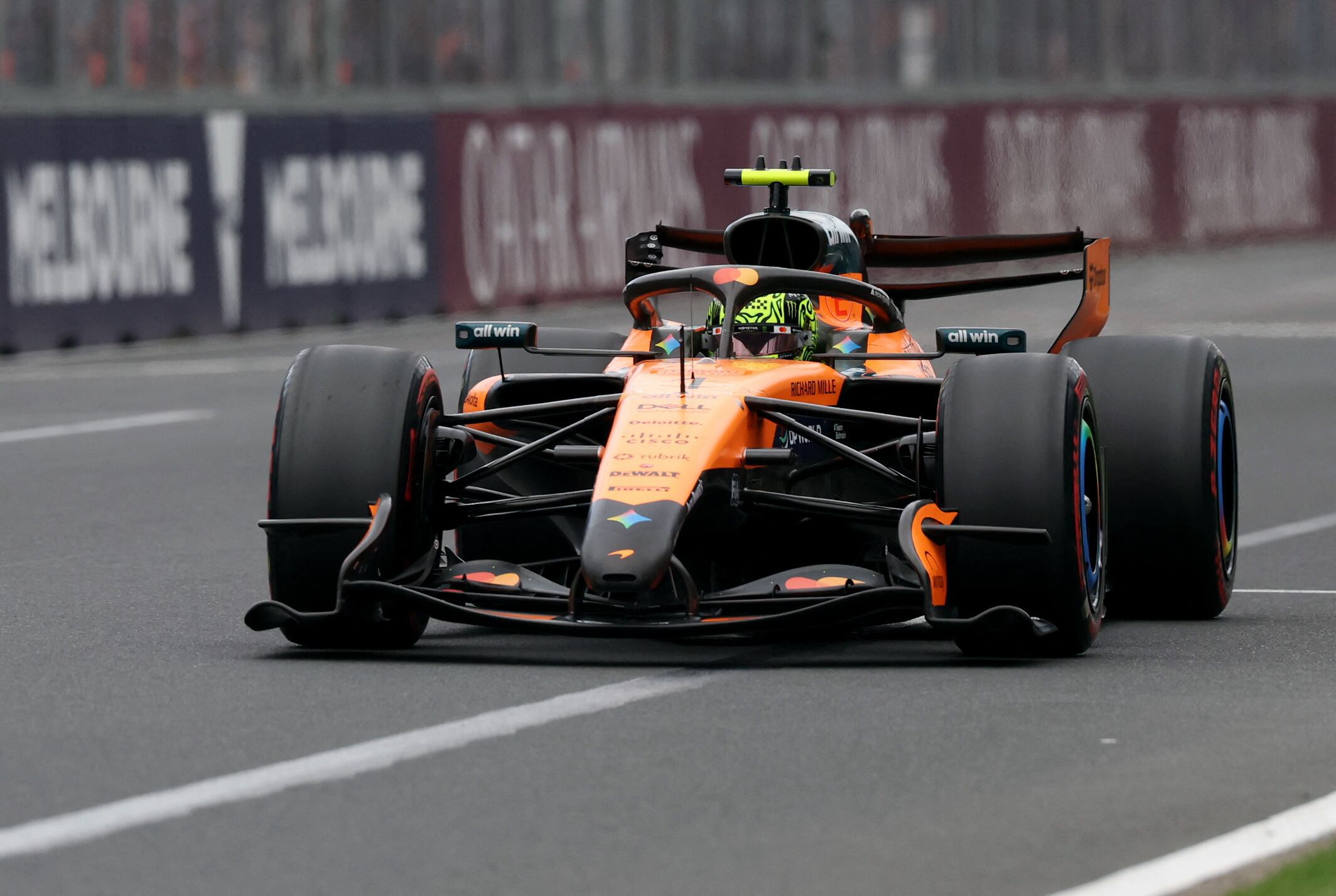 Carro de Fórmula 1 da equipe McLaren, em laranja e preto, correndo em alta velocidade em um circuito urbano, com placas de ‘Melbourne’ ao fundo