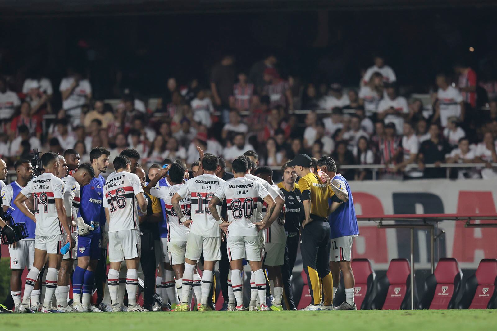 Jogadores do São Paulo reunidos na beira do campo, com torcida ao fundo