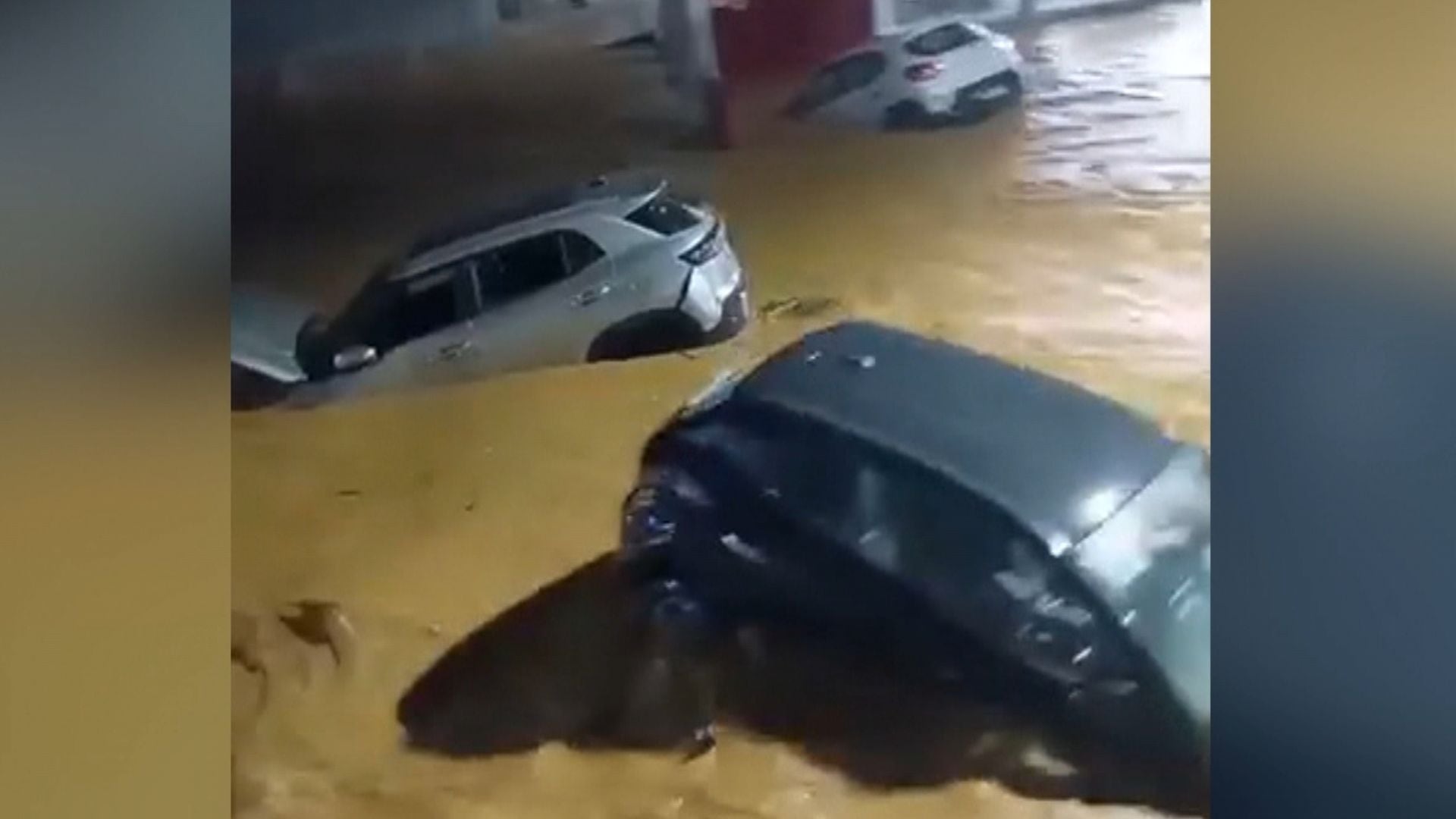Área alagada durante a noite mostra vários carros estão parcialmente submersos em água barrenta, com apenas partes das carrocerias visíveis acima do nível da enchente. O ambiente ao fundo tem estruturas iluminadas, sugerindo que o local é urbano ou parte de um estacionamento coberto.