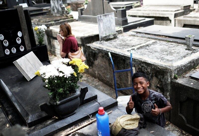 O garoto Gabriel, de 11 anos de idade, sorri e cumprimenta o fotógrafo perto de mulher depois de limpar túmulo em cemitério do Rio de Janeiro nesta quarta-feira (1º). A prática, porém, configura trabalho infantil e será alvo de uma fiscalização das superintendências do Ministério do Trabalho nesta quinta-feira (2)
