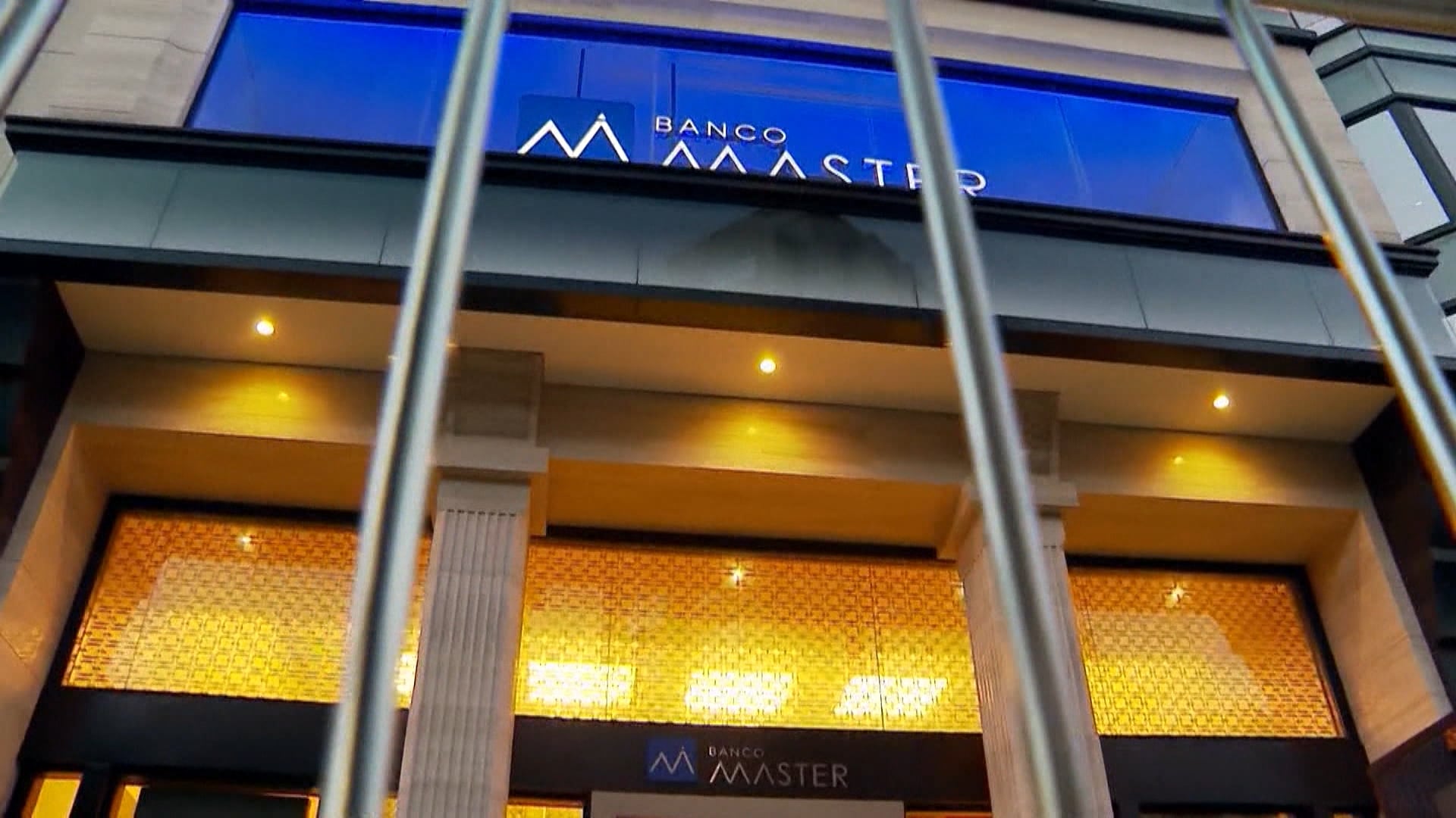 Banco Master com o logo em azul em branco ao centro do prédio, com colunas gregas e luzes amarelas. Em frente ao prédio estão grades fechando o banco.