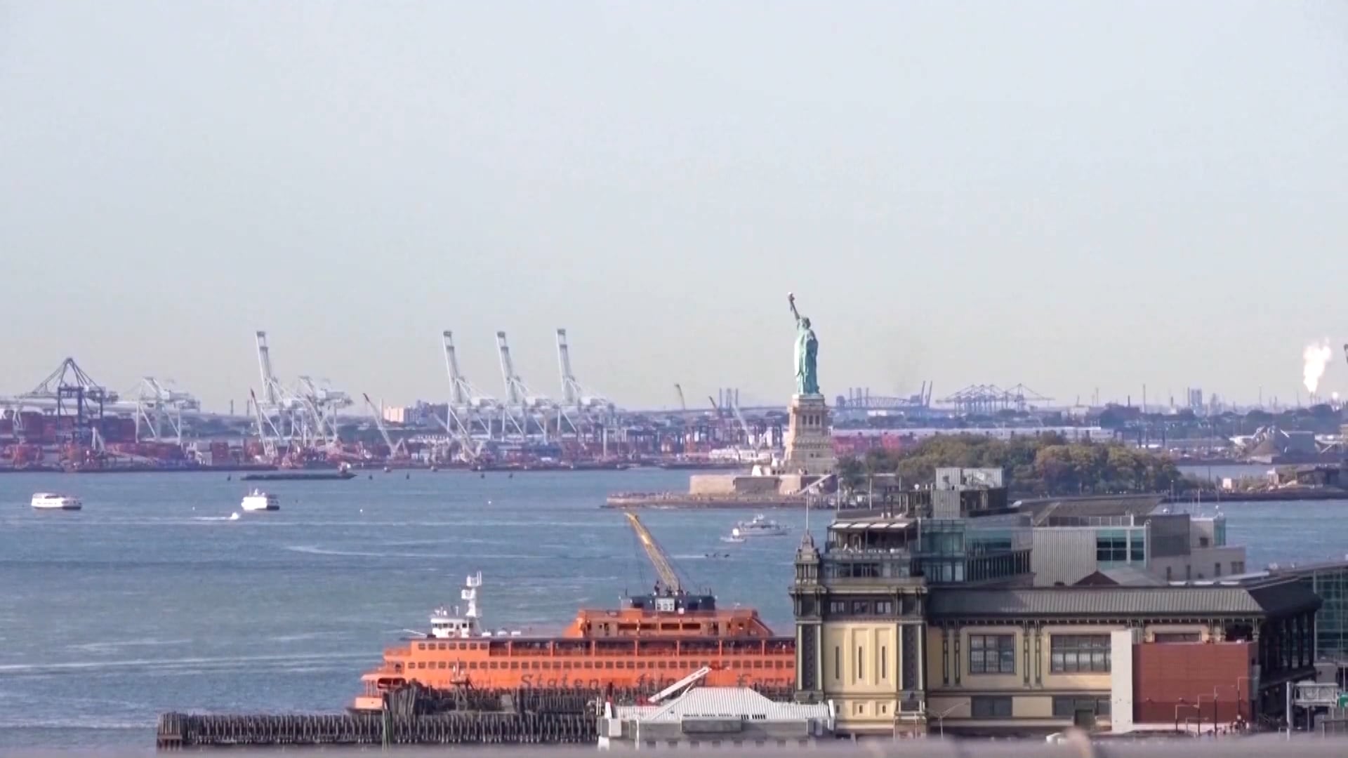 Imagem da Estátua da Liberdade vista à distância, no porto de Nova York. Em primeiro plano, aparece um barco laranja navegando pela água e um prédio. Ao fundo, há guindastes e estruturas portuárias.