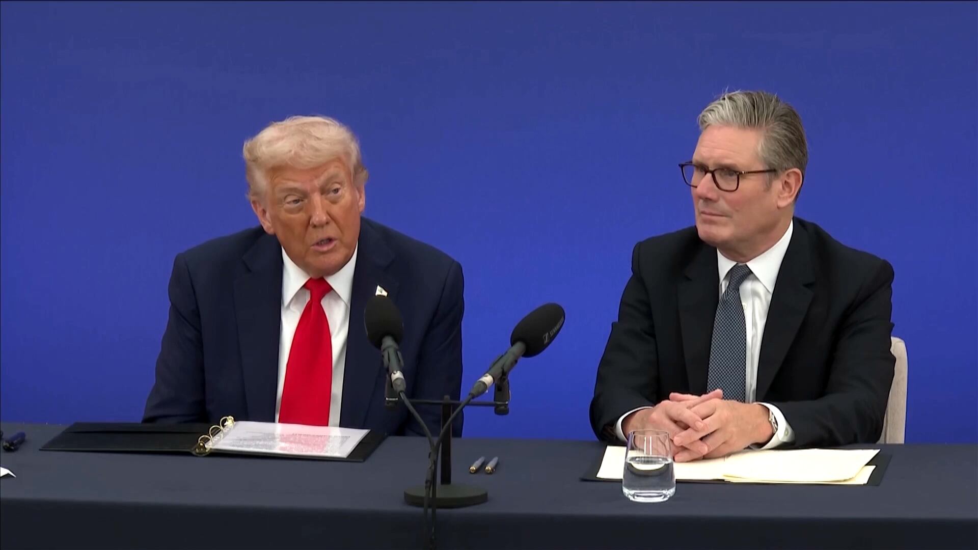 Donald Trump e Keir Starmer sentados lado a lado em uma mesa de conferência, diante de microfones, papéis e um copo de água, com um fundo azul sólido