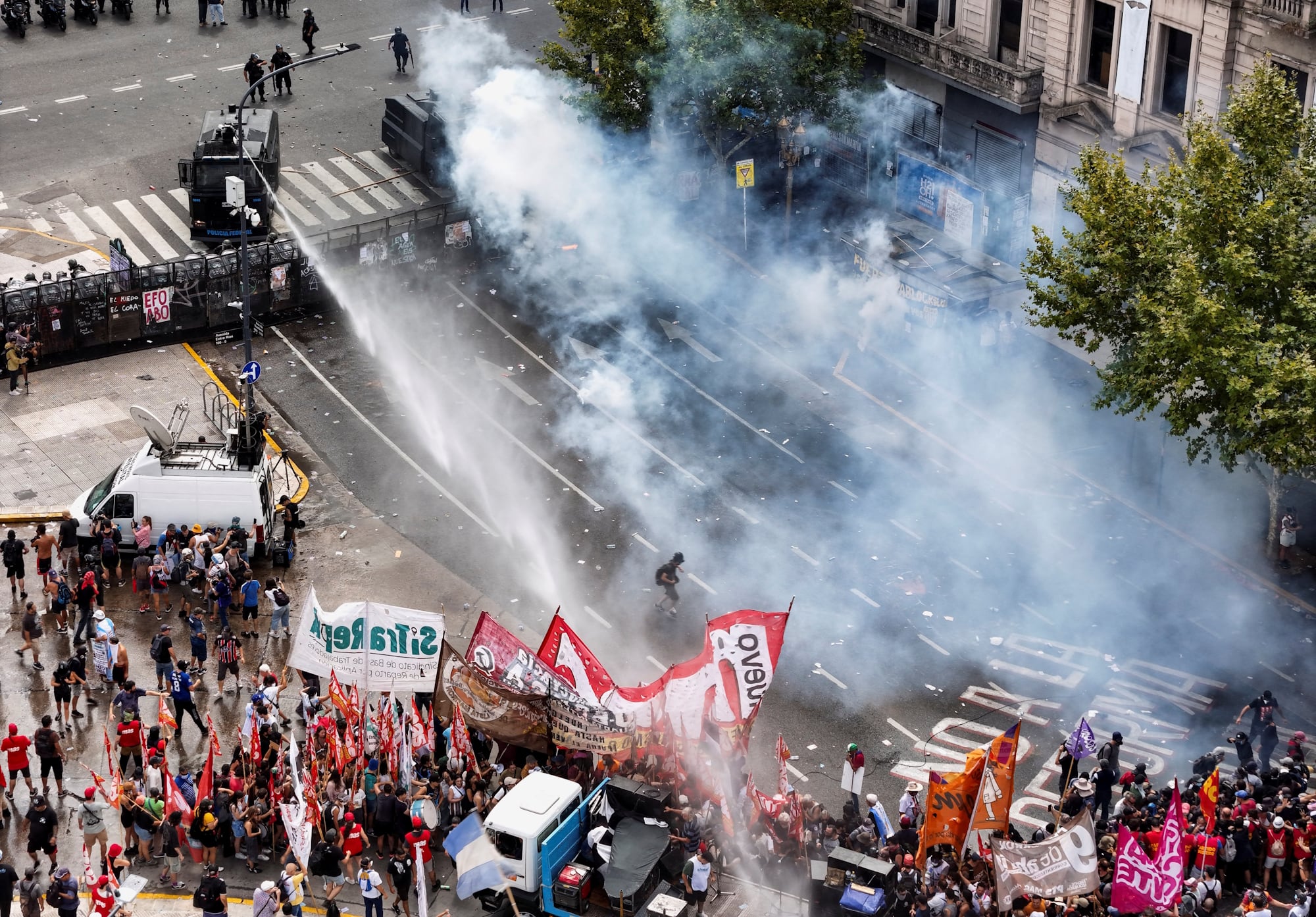 Imagem aérea mostra polícia disparando jatos d'água contra manifestantes, separados por uma cerca, em uma avenida