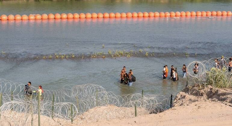 Migrantes caminham entre arame farpado e uma série de boias colocadas nas águas do rio Grande ao longo da fronteira com o México, em Eagle Pass