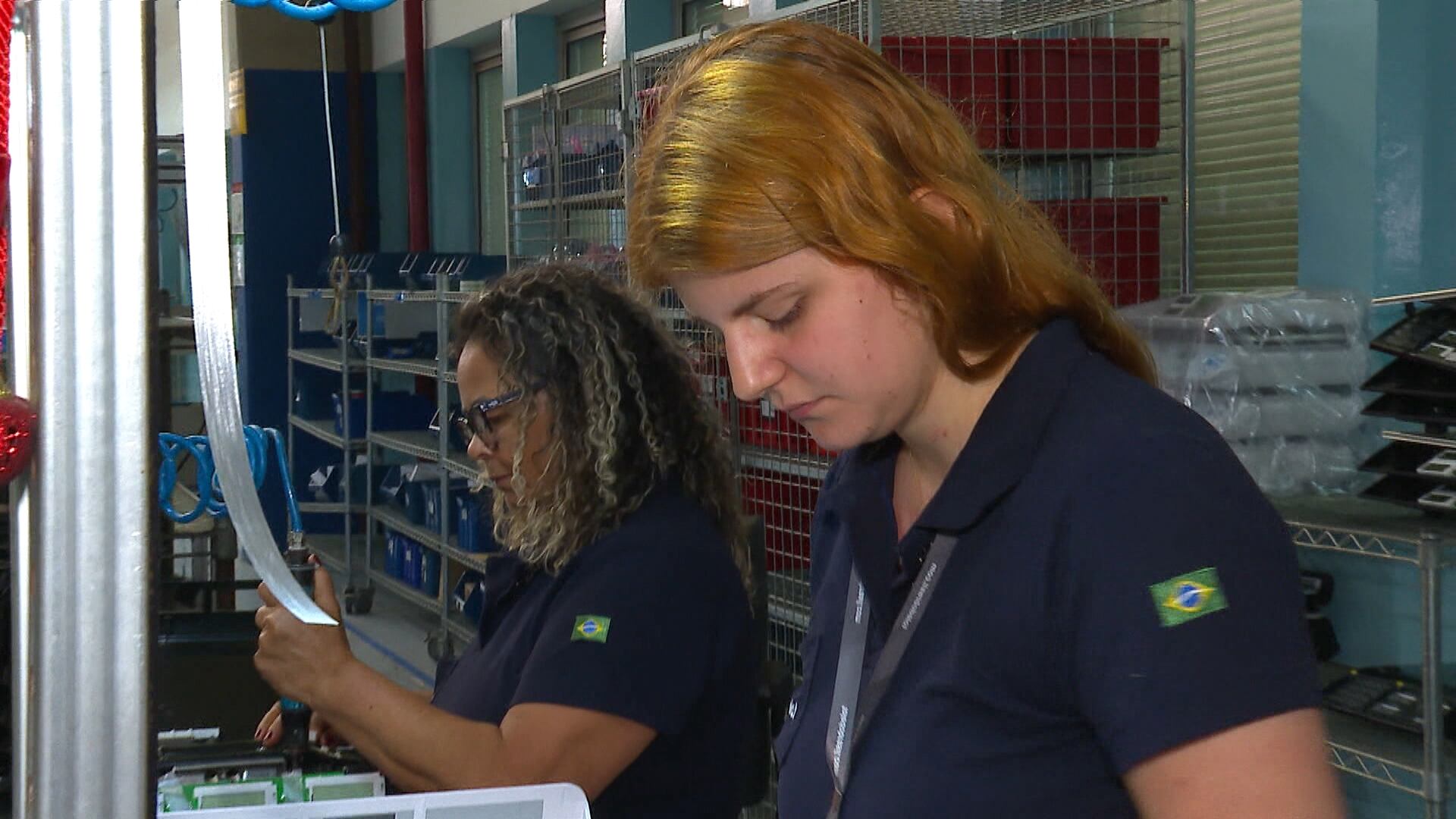 A imagem mostra duas mulheres trabalhando. Ambas vestem camisas escuras com um pequeno emblema da bandeira do Brasil em uma das mangas. Uma das pessoas segura uma folha de papel, enquanto a outra observa objetos sobre uma bancada. O local tem iluminação artificial e diversos itens organizados nas prateleiras ao fundo.