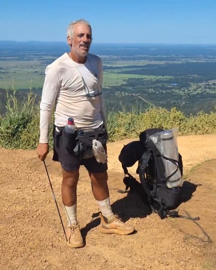 Um homem de cabelos grisalhos, com uma camiseta de manga longa branca e shorts escuros, está em pé em um caminho de terra, segurando um bastão de caminhada. Ele usa meias claras e botas de trekking. Ao seu lado, há uma mochila preta com um compartimento transparente. Ao fundo, uma vasta paisagem verde se estende sob um céu azul claro, com algumas nuvens brancas. A cena transmite uma sensação de aventura ao ar livre.
