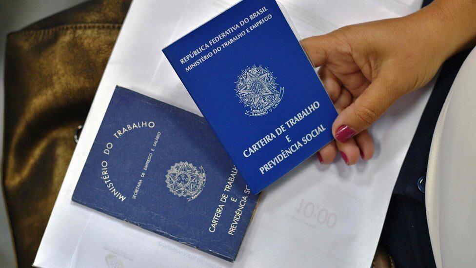 Foco aproximado mostra duas carteiras de trabalho, uma delas segurada por uma mão