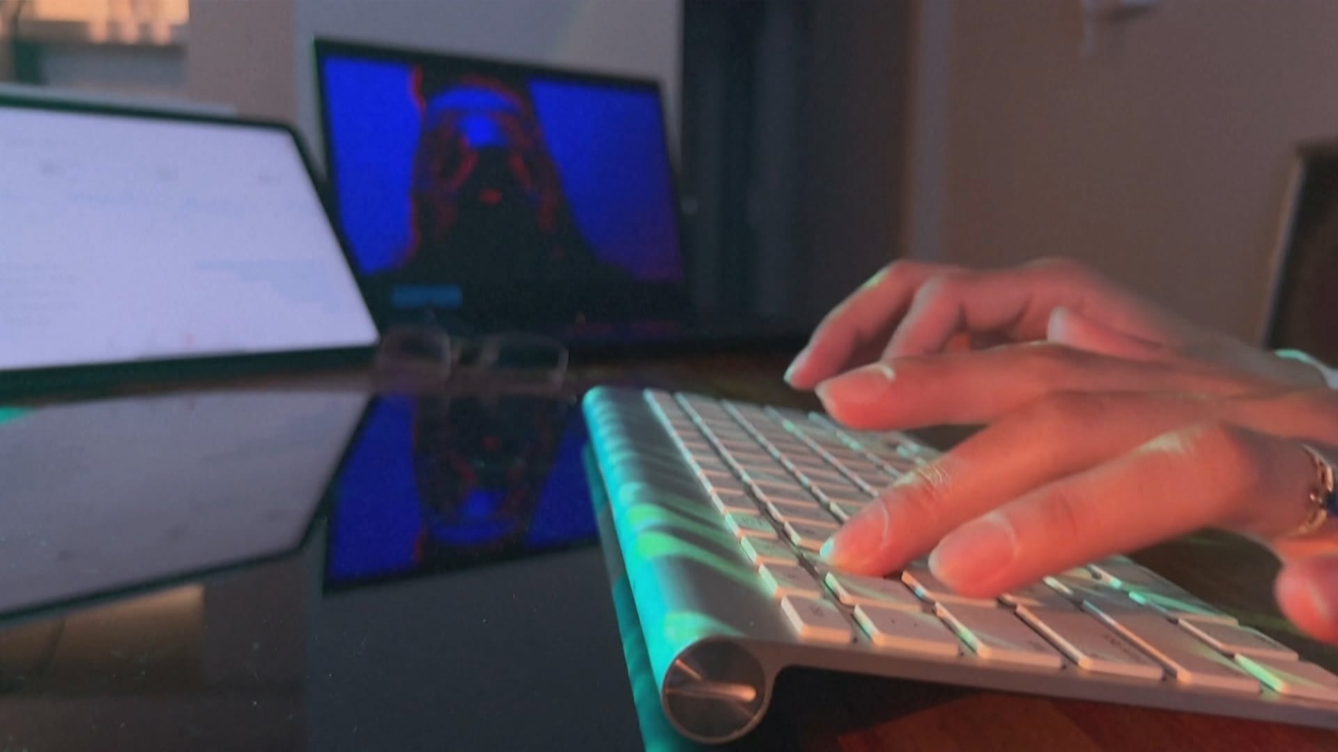 A imagem mostra uma pessoa digitando em um teclado branco sobre uma superfície brilhante. Ao fundo, há duas telas: uma exibindo uma figura iluminada em tons de azul e vermelho, e outra com conteúdo claro, parcialmente fora de foco.
