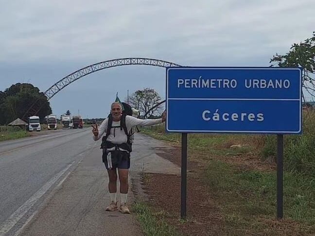 Um homem de mochila, com roupas leves e sandálias, está parado ao lado de uma placa azul que indica "PERÍMETRO URBANO - Cáceres". Ao fundo, há um arco metálico e uma estrada com alguns veículos passando. O céu está nublado, criando uma atmosfera tranquila. O cenário é rural, com vegetação à beira da estrada.