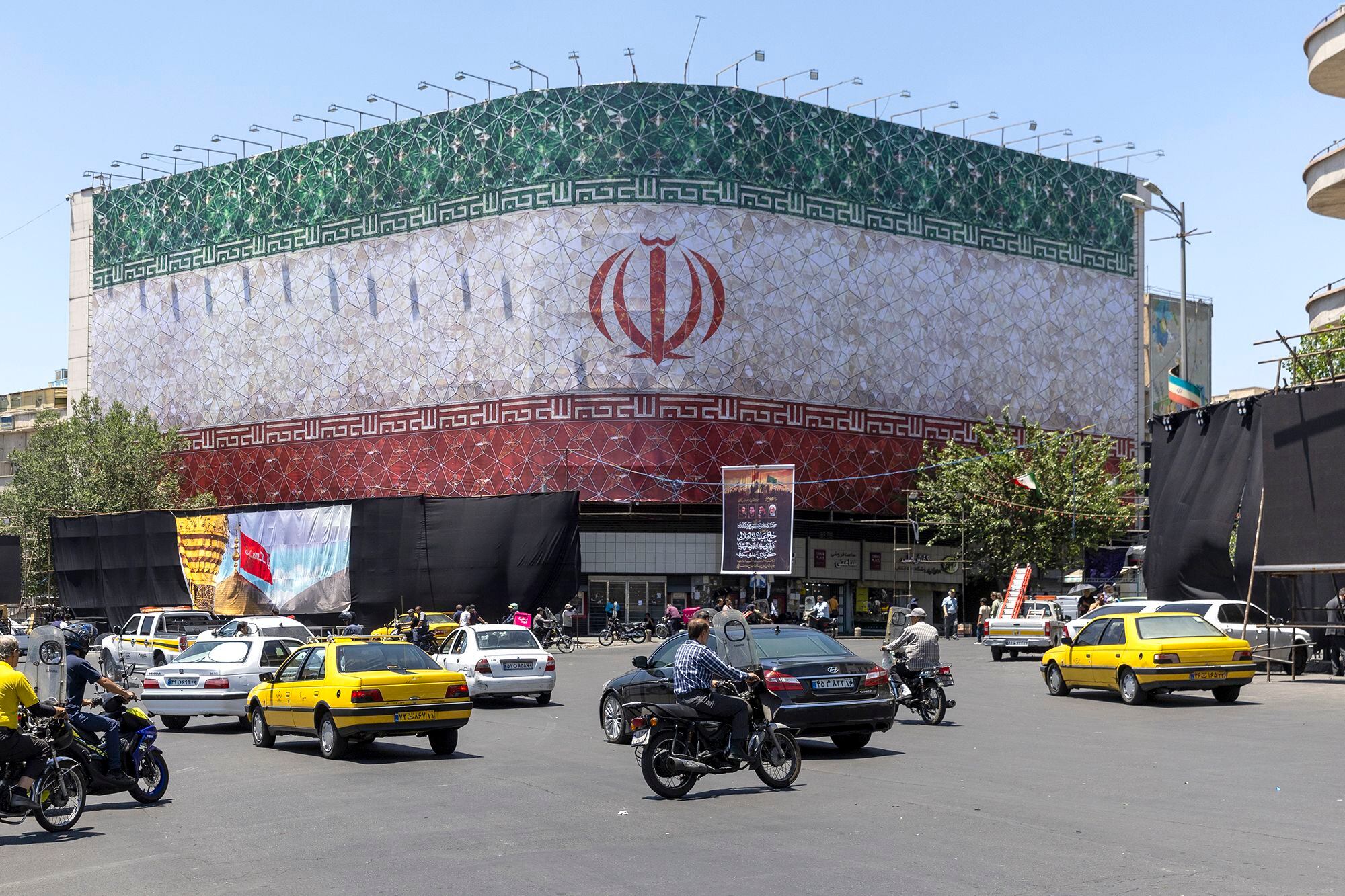 Carros e monociclos são vistos circulando em frente a um outdoor com a bandeira iraniana em Teerã