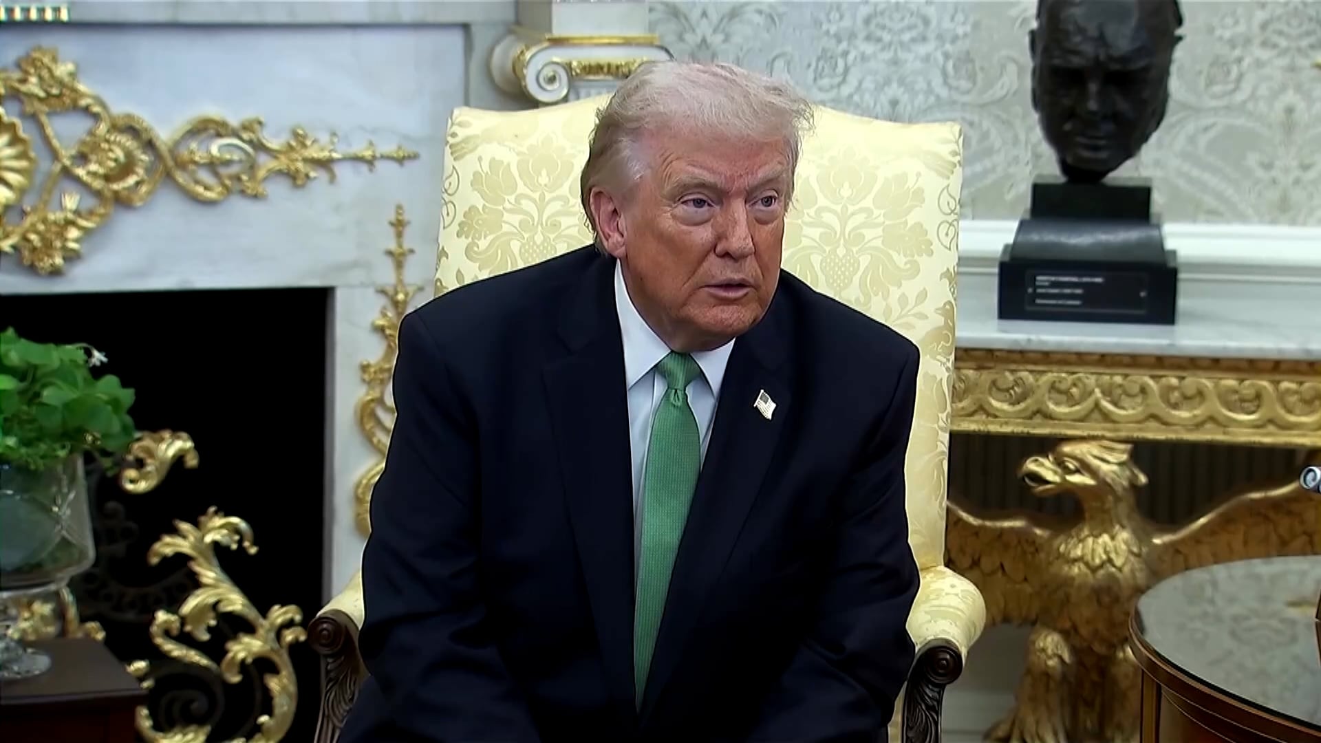 Donald Trump sentado em uma poltrona clara dentro de uma sala decorada com móveis elegantes e ornamentos dourados, vestindo um terno escuro e gravata verde