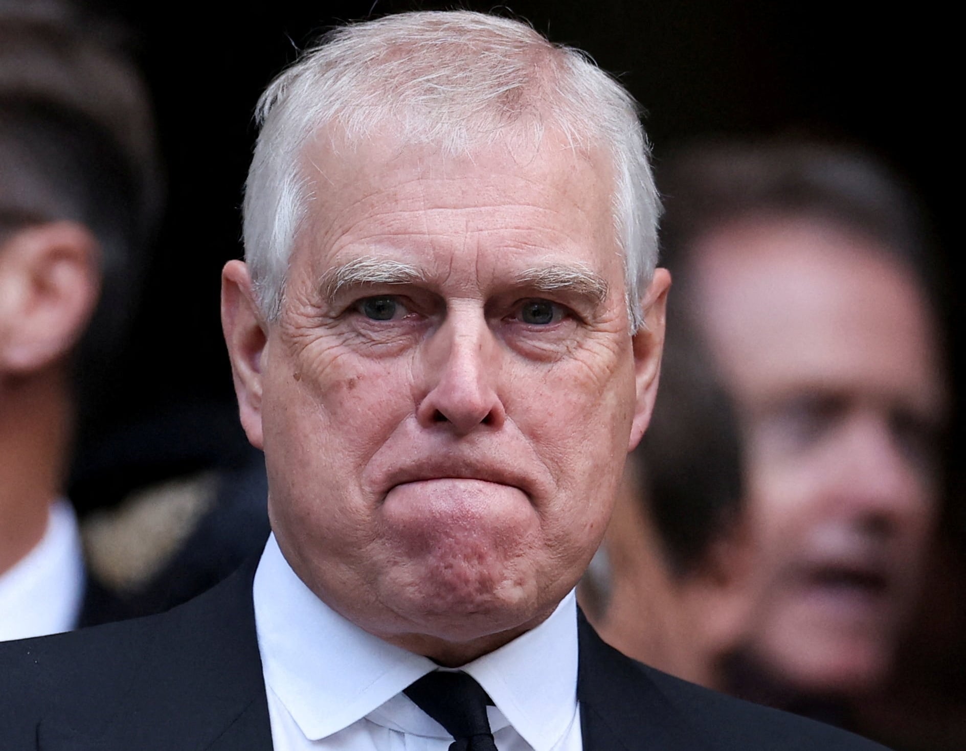 Homem grisalho, de terno, expressa tristeza com os músculos do rosto com pessoas ao fundo em segundo plano