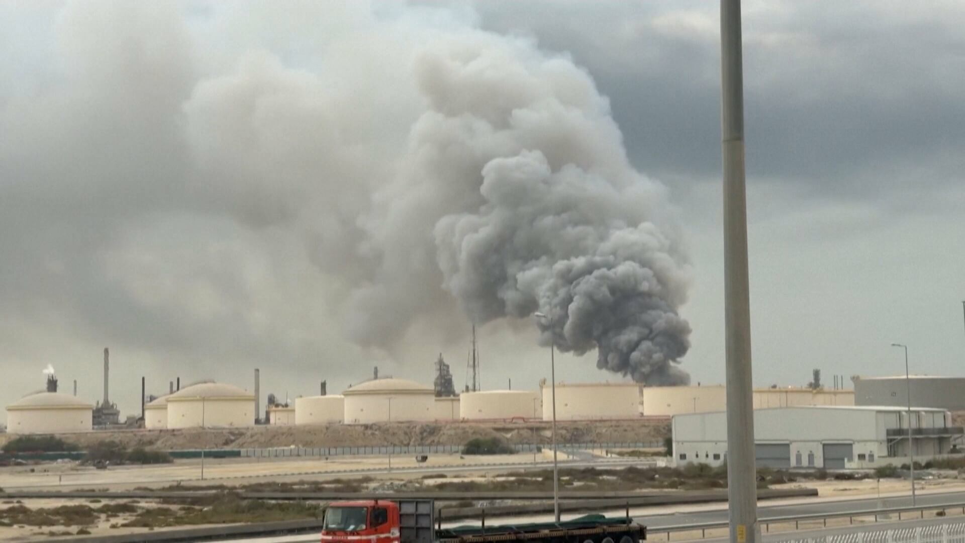 Complexo industrial com tanques cilíndricos de armazenamento, de onde surge uma densa nuvem de fumaça cinza escura elevando‑se ao céu, indicando um possível incêndio ou explosão