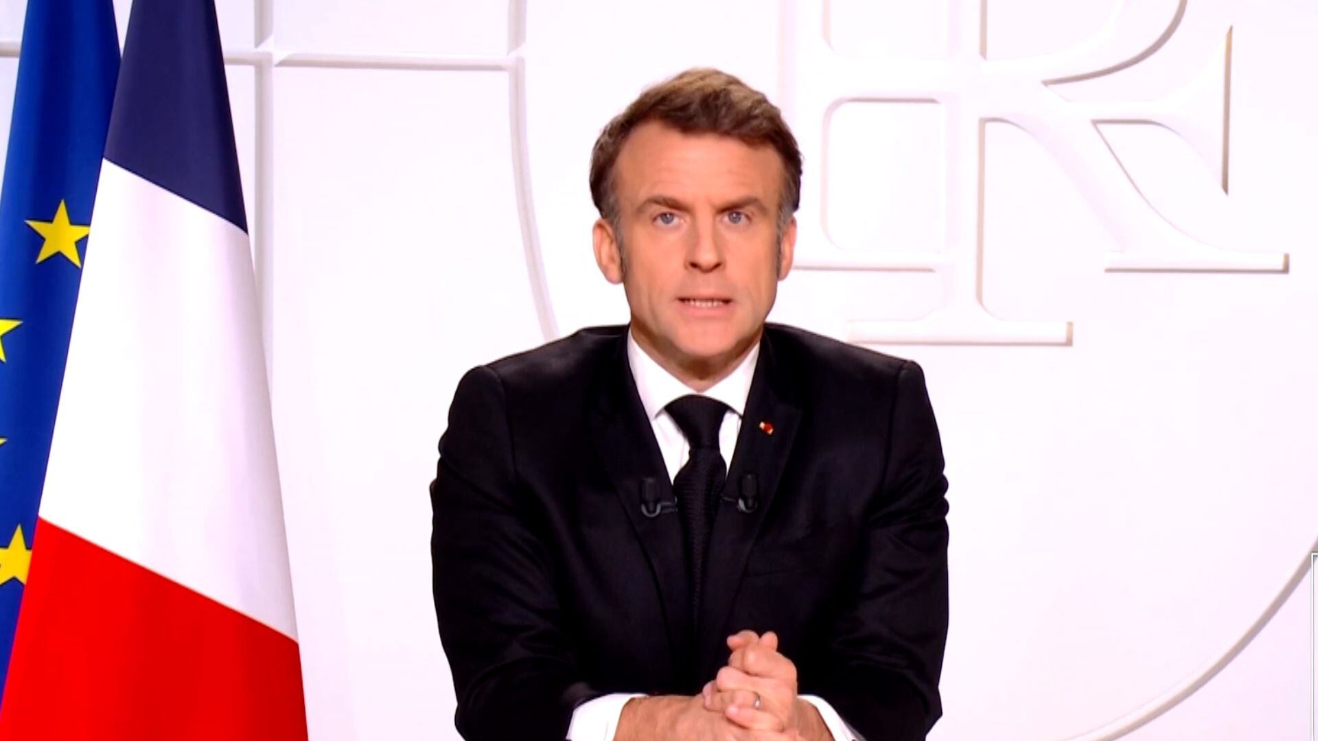 Macron ao centro, com terno e gravata preta e camisa branca, está com postura séria. Na esquerda da imagem estão colocadas as bandeiras da França e da União Europeia