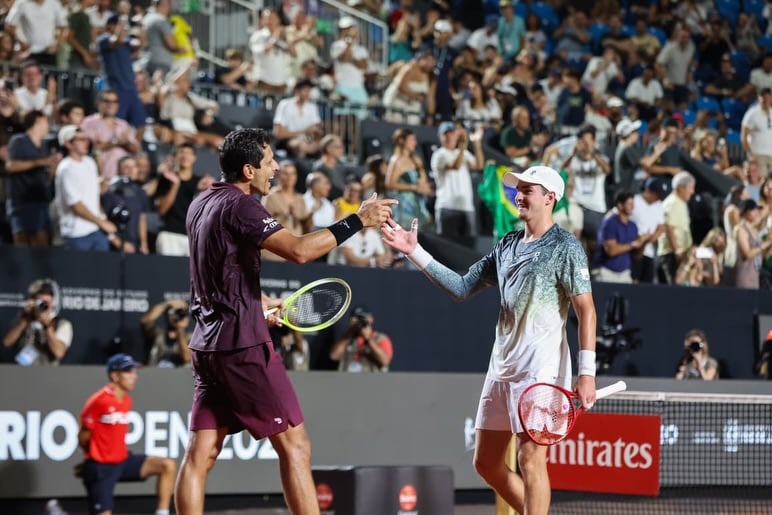 Com uma arquibancada cheia ao fundo, Marcelo Melo, vestido shorts e camiseta vinho, cumprimenta João Fonseca, com short branco e camiseta branca na base e com degradê verde e amarelo na parte de cima.