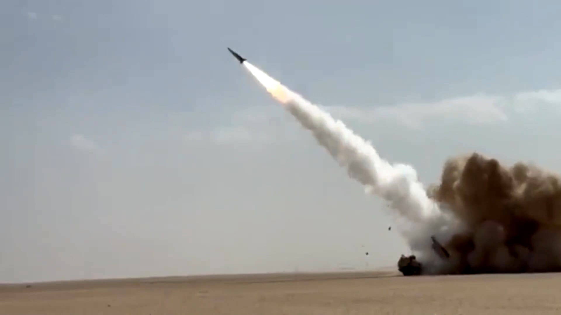A imagem mostra um míssil sendo lançado em uma área desértica aberta. Ele aparece inclinado para cima, deixando um rastro branco de fumaça enquanto sobe. No solo, há um veículo militar parcialmente envolto por poeira e fumaça levantadas pelo lançamento. O céu está claro e sem nuvens.