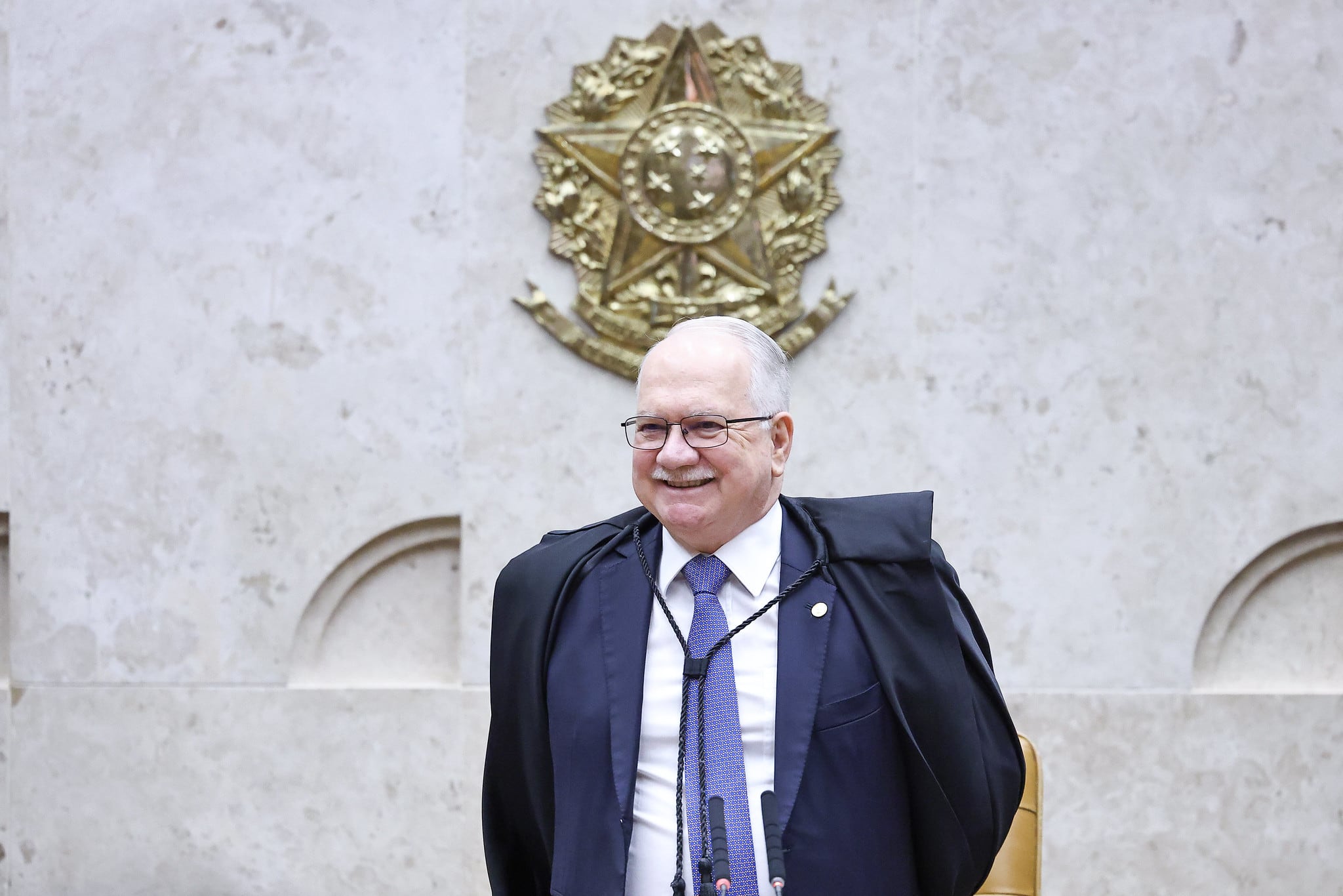 Ministro Edson Fachin veste um terno e sorri. Ele está em pé em frente ao brazão do Brasil