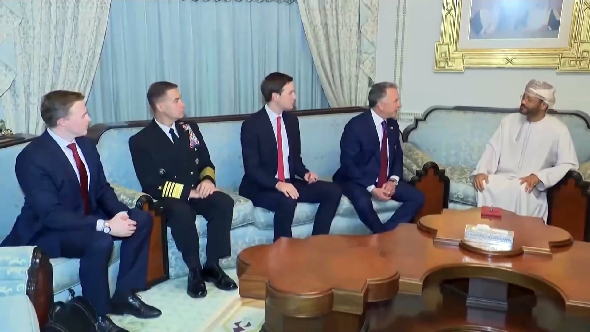 Reunião formal em uma sala mobiliada com sofás claros e mesa de centro de madeira, onde cinco pessoas sentadas conversam em um ambiente decorado com molduras e cortinas
