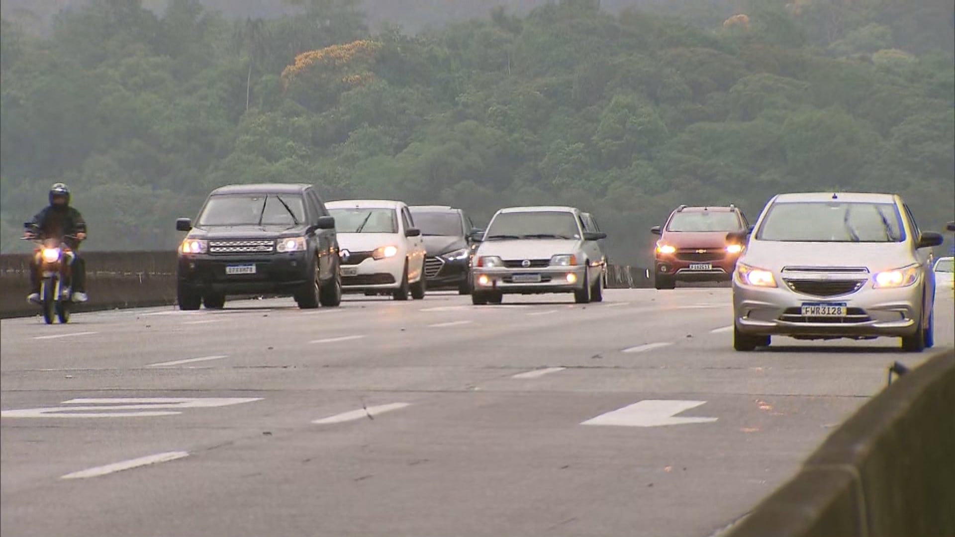 Carros e uma motocicleta trafegam por uma rodovia larga em um dia nublado, com vegetação densa ao fundo