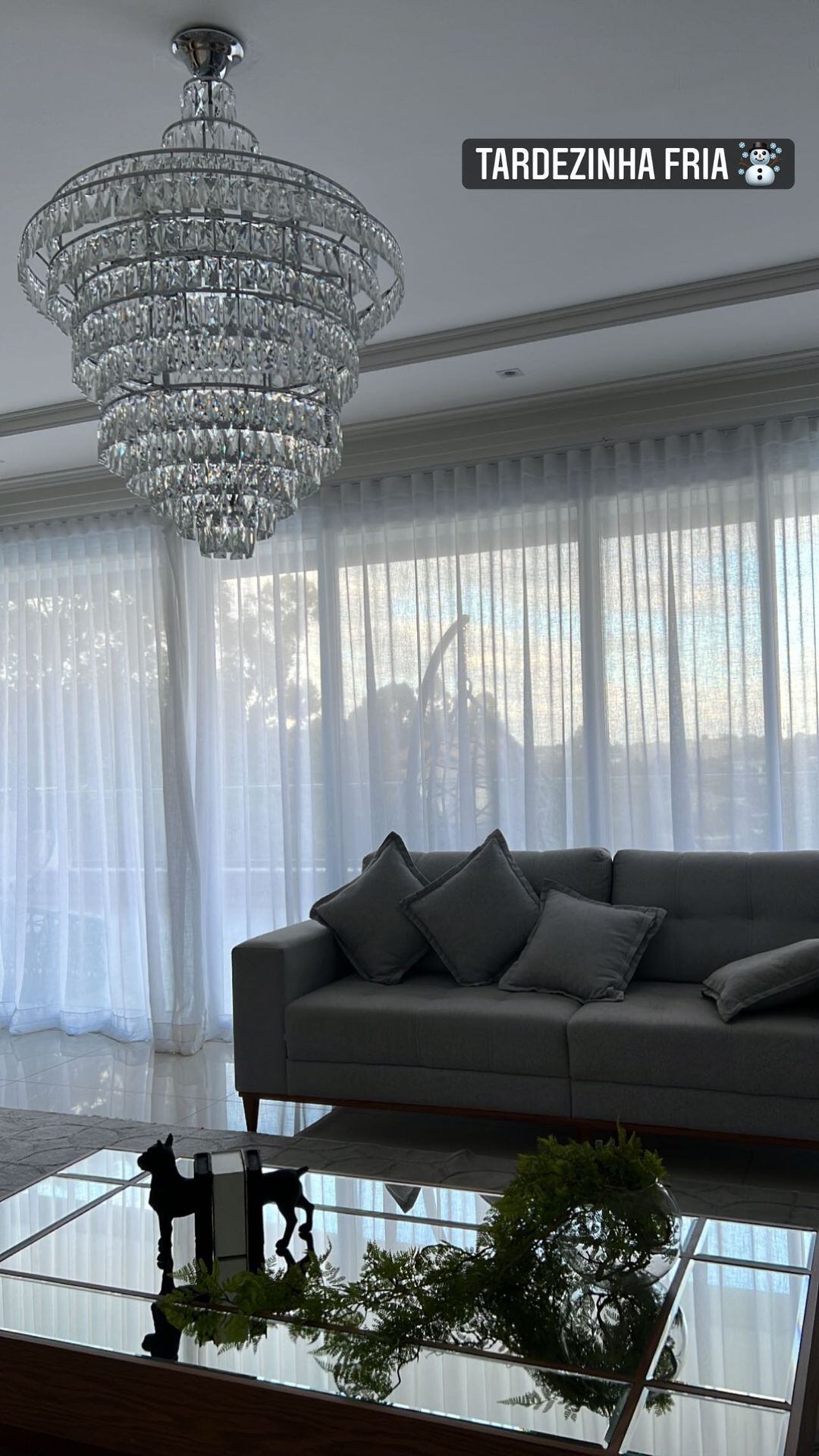 A sala tem um lustre de cristal, sofás em tons de cinza e uma mesa de centro espelhada