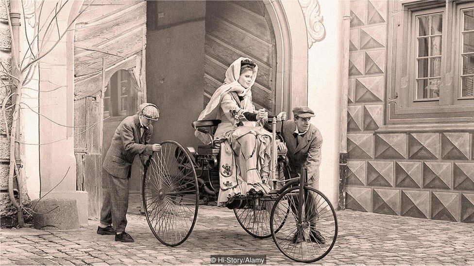 Bertha Benz sobre seu carro, com rodas de madeira, ao lado de Karl Benz