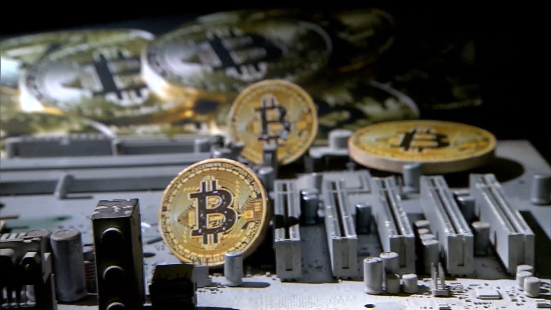 Várias moedas douradas com o símbolo do Bitcoin posicionadas sobre uma superfície, que parece uma placa eletrônica ou circuito. O fundo está desfocado, com outras moedas semelhantes visíveis.