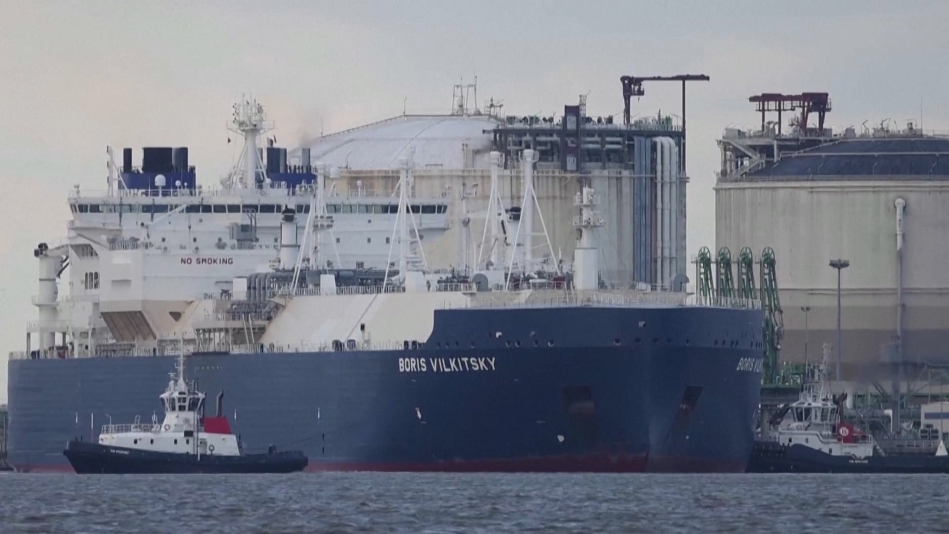 A imagem mostra um grande navio cargueiro azul-escuro com o nome “BORIS VILKITSKY” pintado na parte frontal. A embarcação está ancorada próximo a instalações industriais costeiras, onde há tanques cilíndricos brancos de grande porte e estruturas metálicas relacionadas ao processamento ou armazenamento de combustíveis. Na água, ao lado do navio maior, há rebocadores menores também em operação. O cenário apresenta céu nublado e o mar calmo.