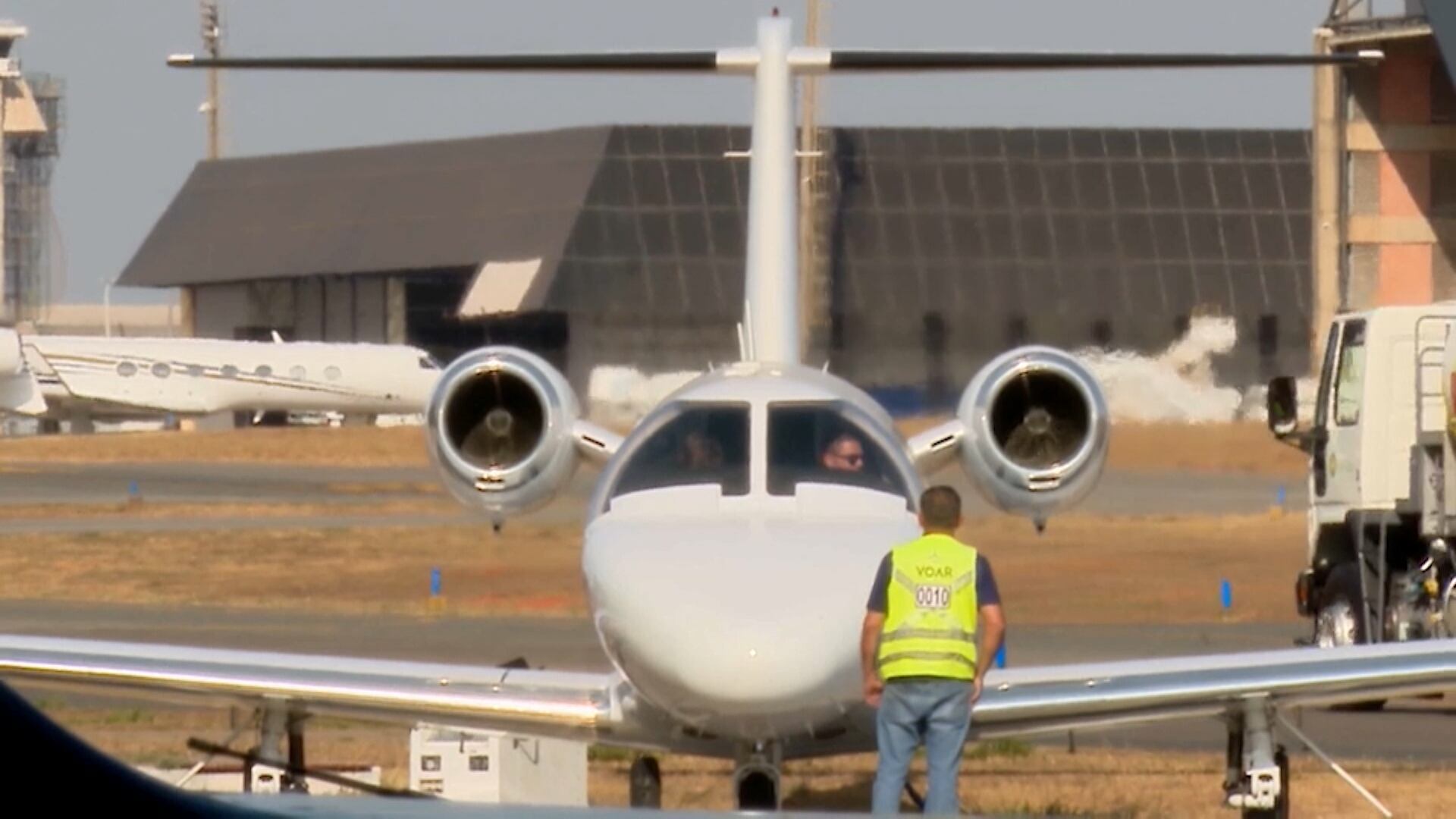 Um jato executivo branco está estacionado em um aeroporto, visto de frente. Um homem, usando um colete amarelo refletivo, está em pé na pista, próximo à aeronave, observando. Ao fundo, é possível ver outras aeronaves e uma estrutura de hangar com uma fachada escura.