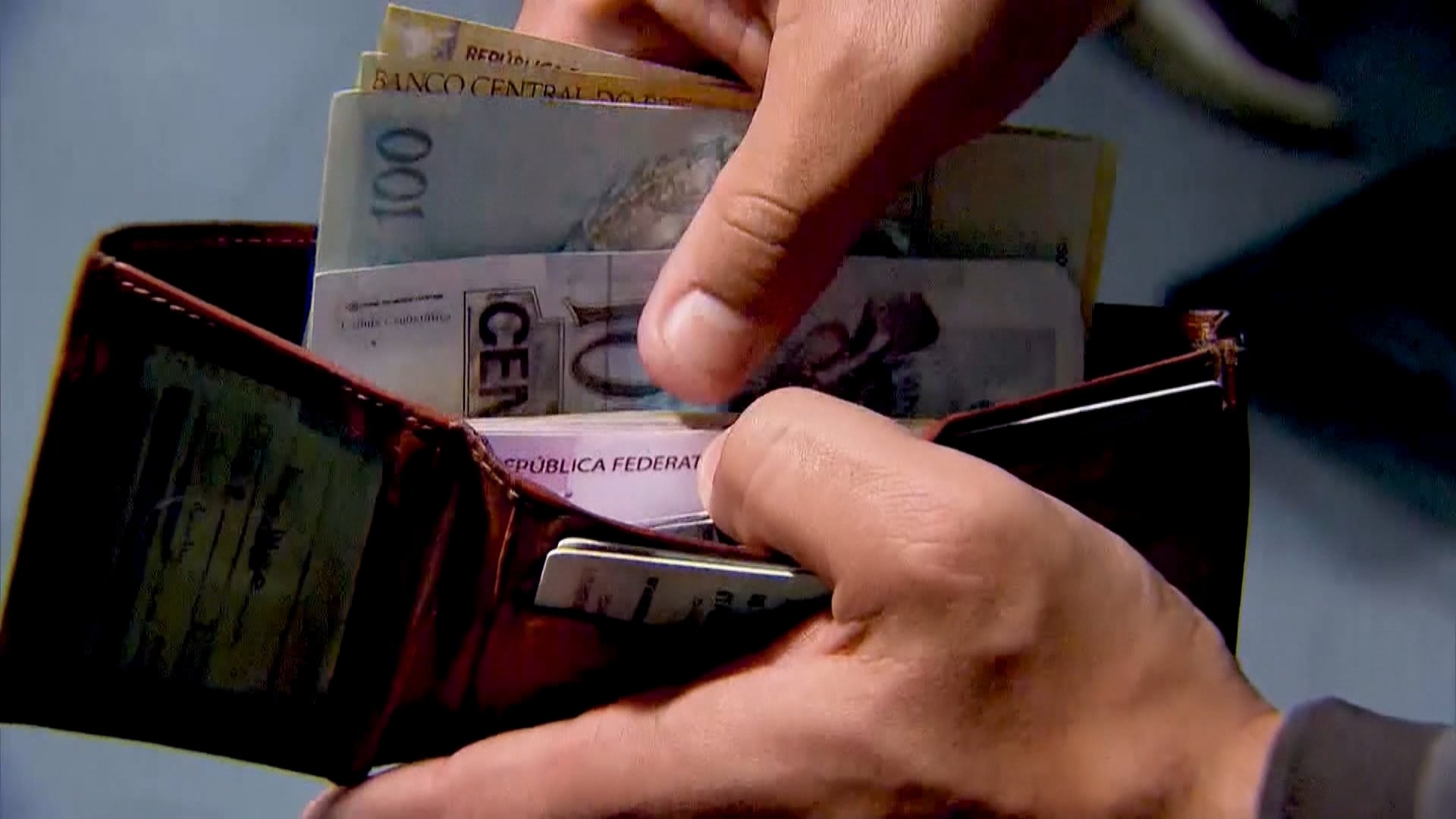 Mãos seguram uma carteira na cor marrom, a esquerda retira de dentro do objeto cédulas de dinheiro com notas de R$ 100 e R$ 50.
