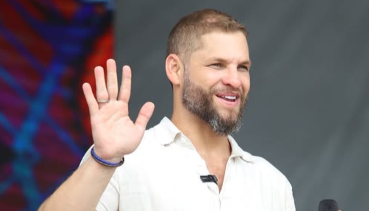 Marcos Cesar, homem branco, de cabelo curto e barba, com uma camiseta branca em um palco de evento